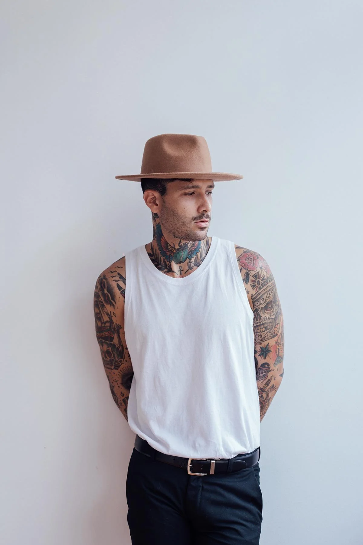 A man with tattoos on his arms and neck, wearing a white sleeveless shirt, black pants, and a tan wide-brimmed hat, standing against a plain white wall.