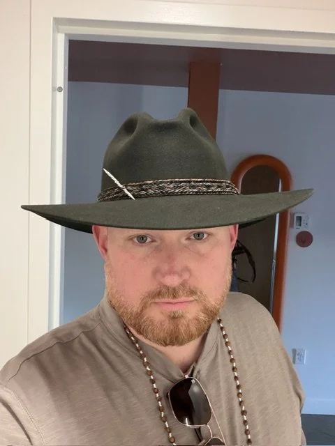 Man wearing a green wide-brimmed hat with a feather, sunglasses hanging from his shirt collar, and a beaded necklace, indoors with a doorway and orange accent in the background.