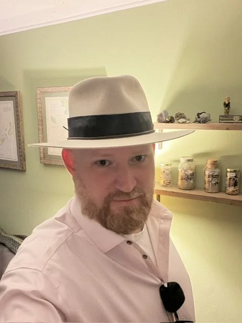 A man with a beard and mustache wearing a white fedora hat with a black band, pink collared shirt, in a room with green walls and shelves holding jars of specimens and rocks.