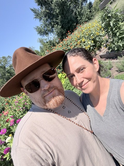A man and a woman taking a selfie outdoors in a garden with colorful flowers and trees.