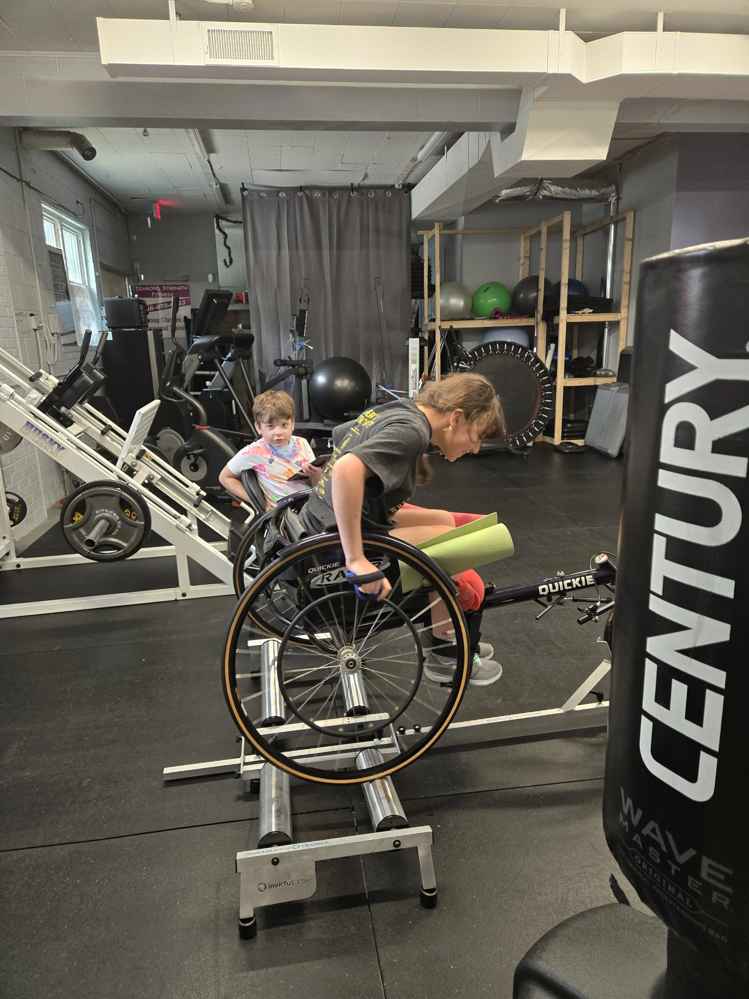 Person in a wheelchair exercising on a rowing machine inside a gym with other fitness equipment and a boy sitting on a nearby machine.