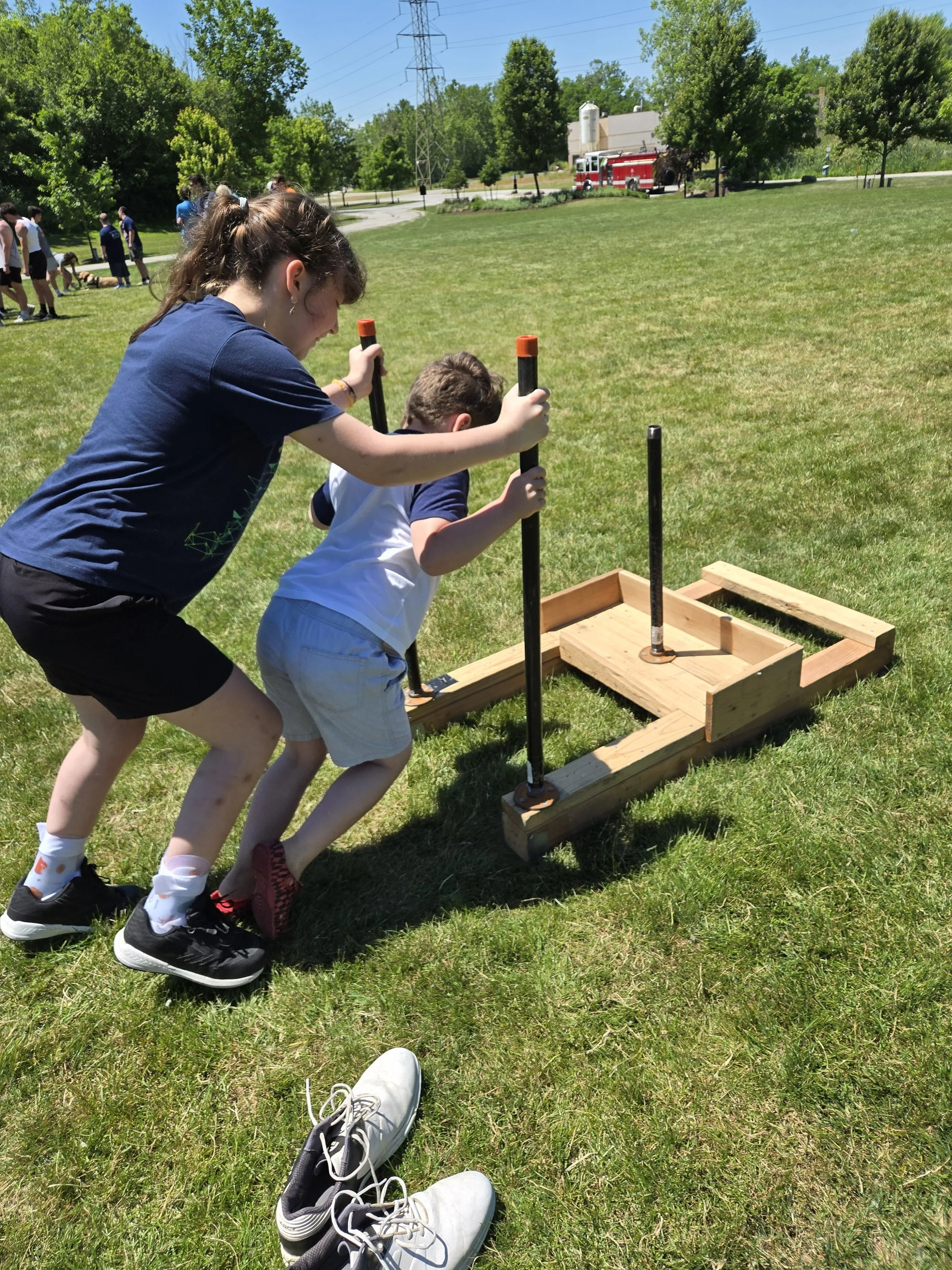 Two children are pushing a wooden box with metal poles in an open grassy field, with other children and a fire truck in the background, trees, and power lines.