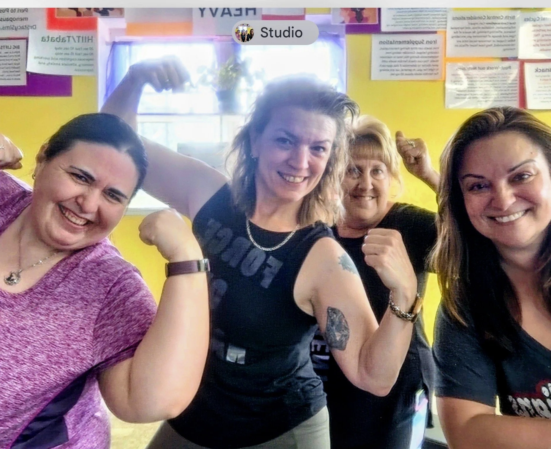 A group of five women posing together, flexing their arms and smiling at the camera inside a colorful room with posters on the yellow wall in the background.