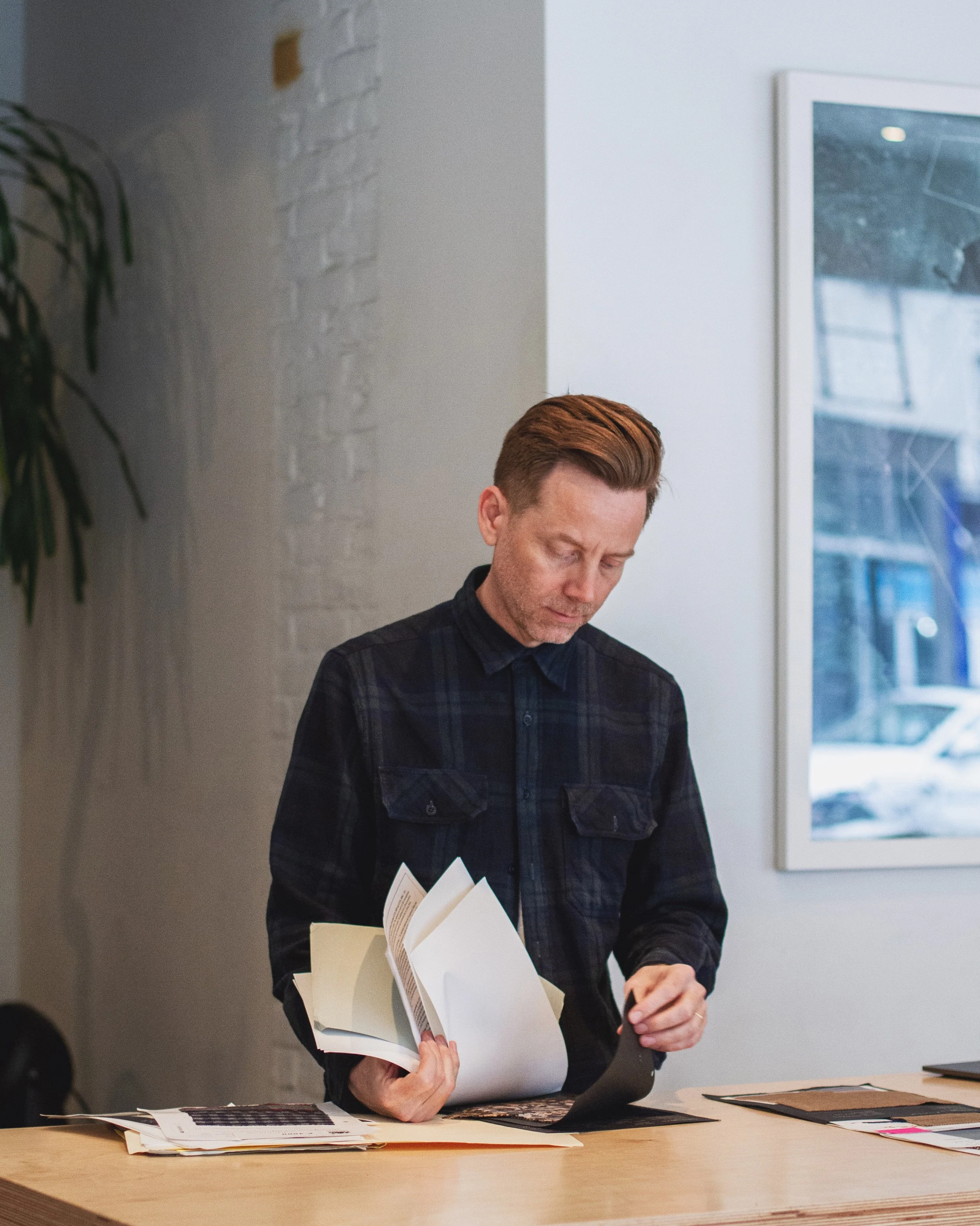 A person with reddish-brown hair is standing at a table, reading a book, with papers and color samples on the table. The person is wearing a dark plaid shirt, and the background features a white wall with framed artwork and a large window.