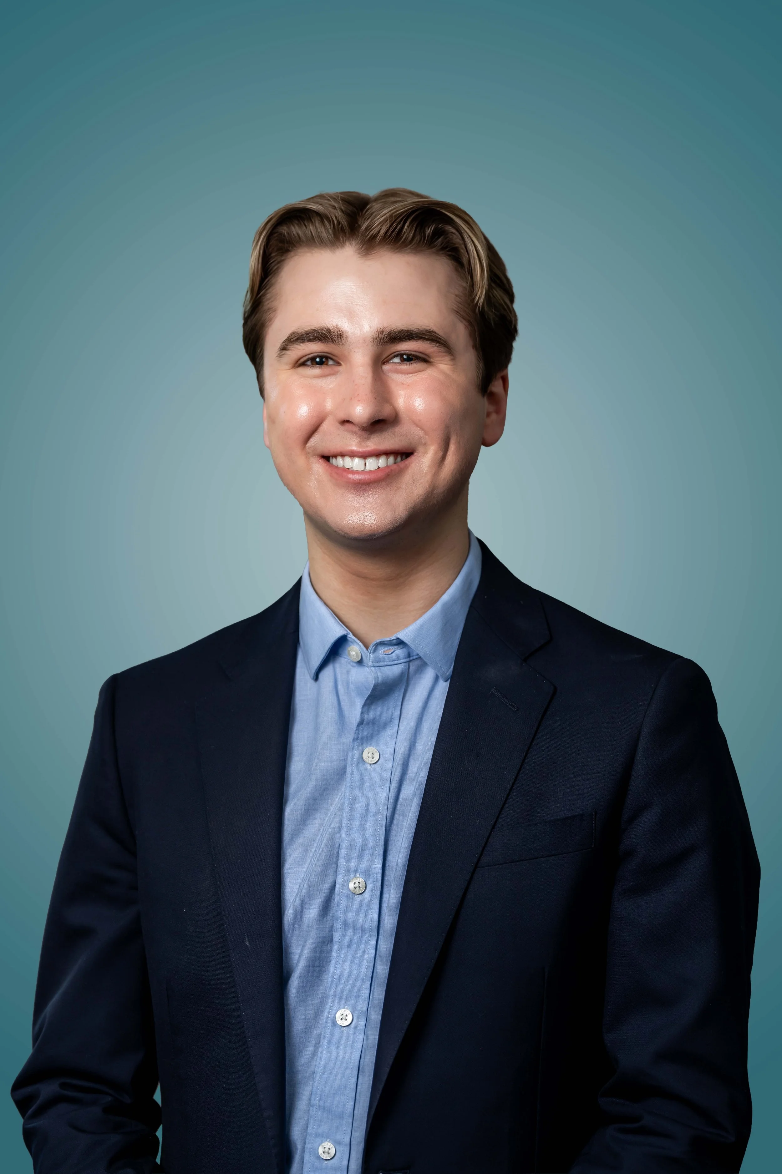Portrait of a young man with brown hair in a professional suit and light blue shirt, smiling, against a teal gradient background.