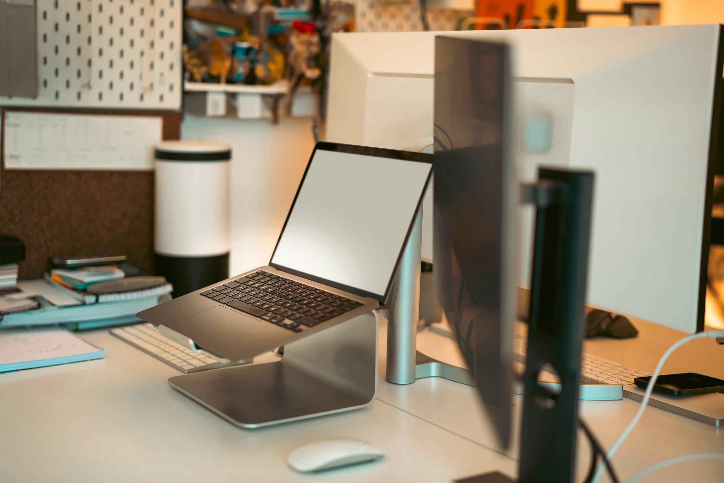 A modern workspace with a MacBook pro laptop placed on a stand, surrounded by books, a tablet, and various office supplies, with a monitor out of focus in the background.