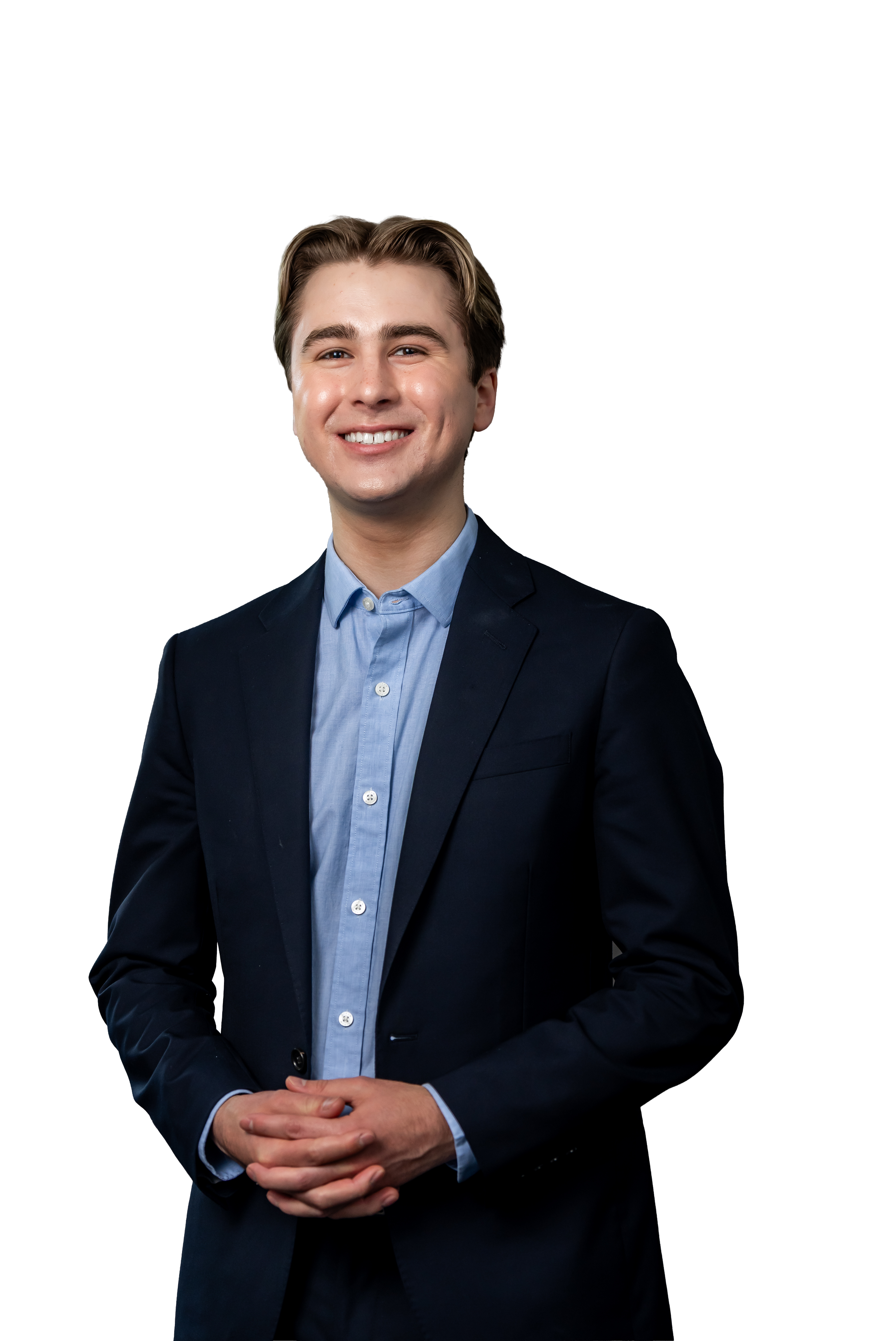 A young man with light skin, brown hair, and a bright smile is wearing a navy blue suit and a light blue dress shirt. He has his hands clasped in front of him and is standing against a plain background.