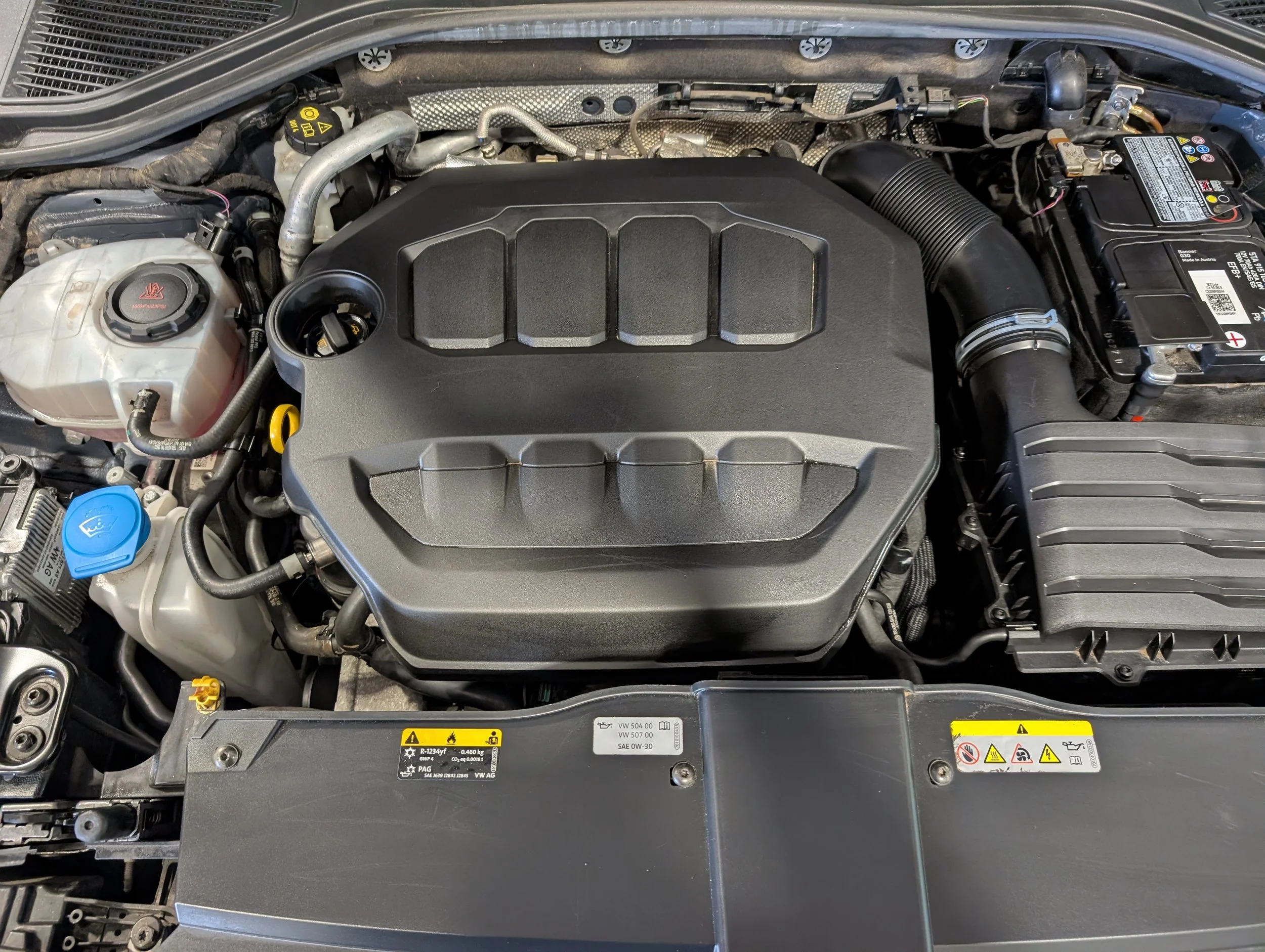 An image of a car engine bay, showing various components such as the oil cap, coolant reservoir, battery, and intake hose.