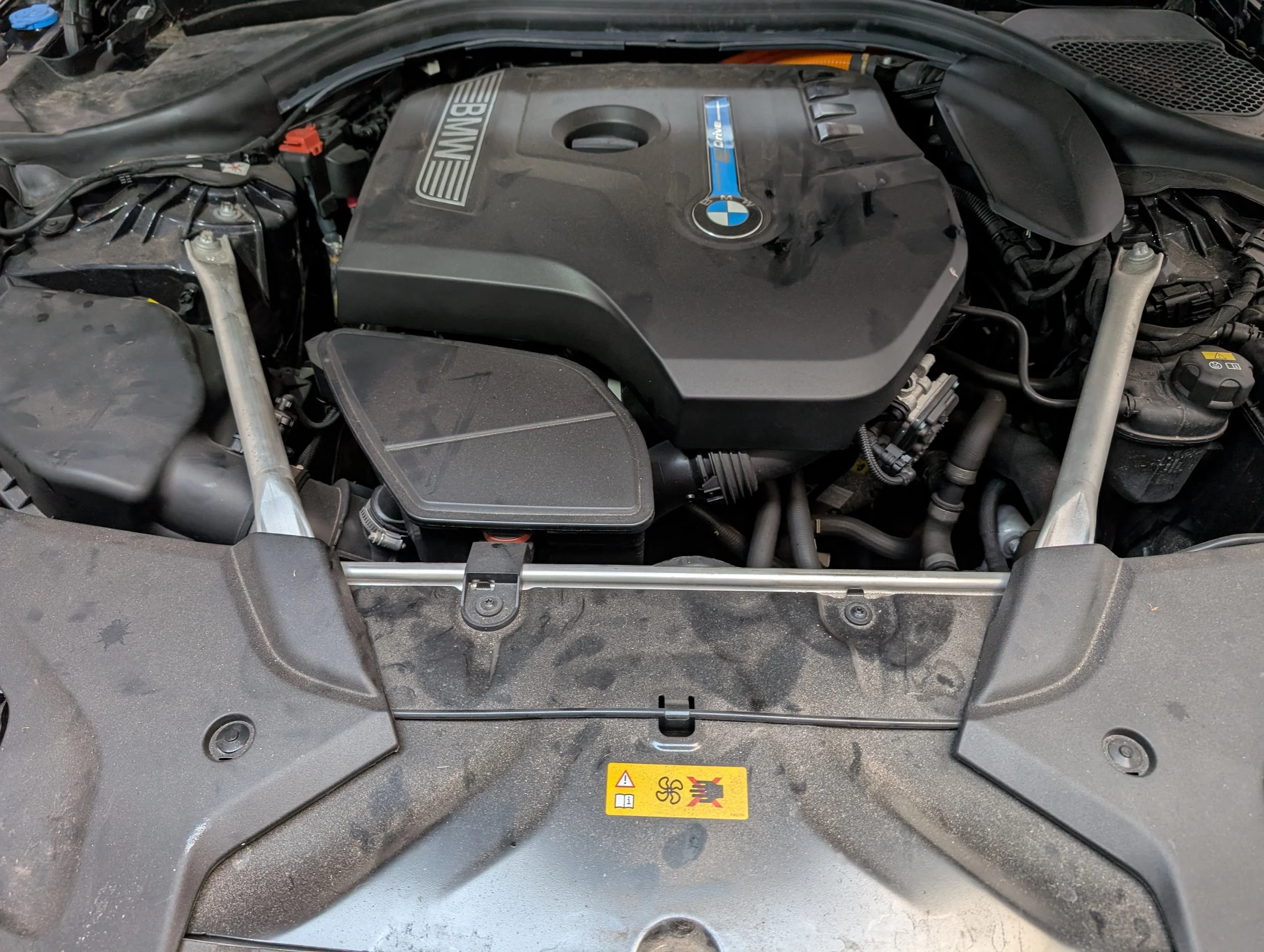 Under the hood of a BMW car showing engine components with a warning label on the lower part.