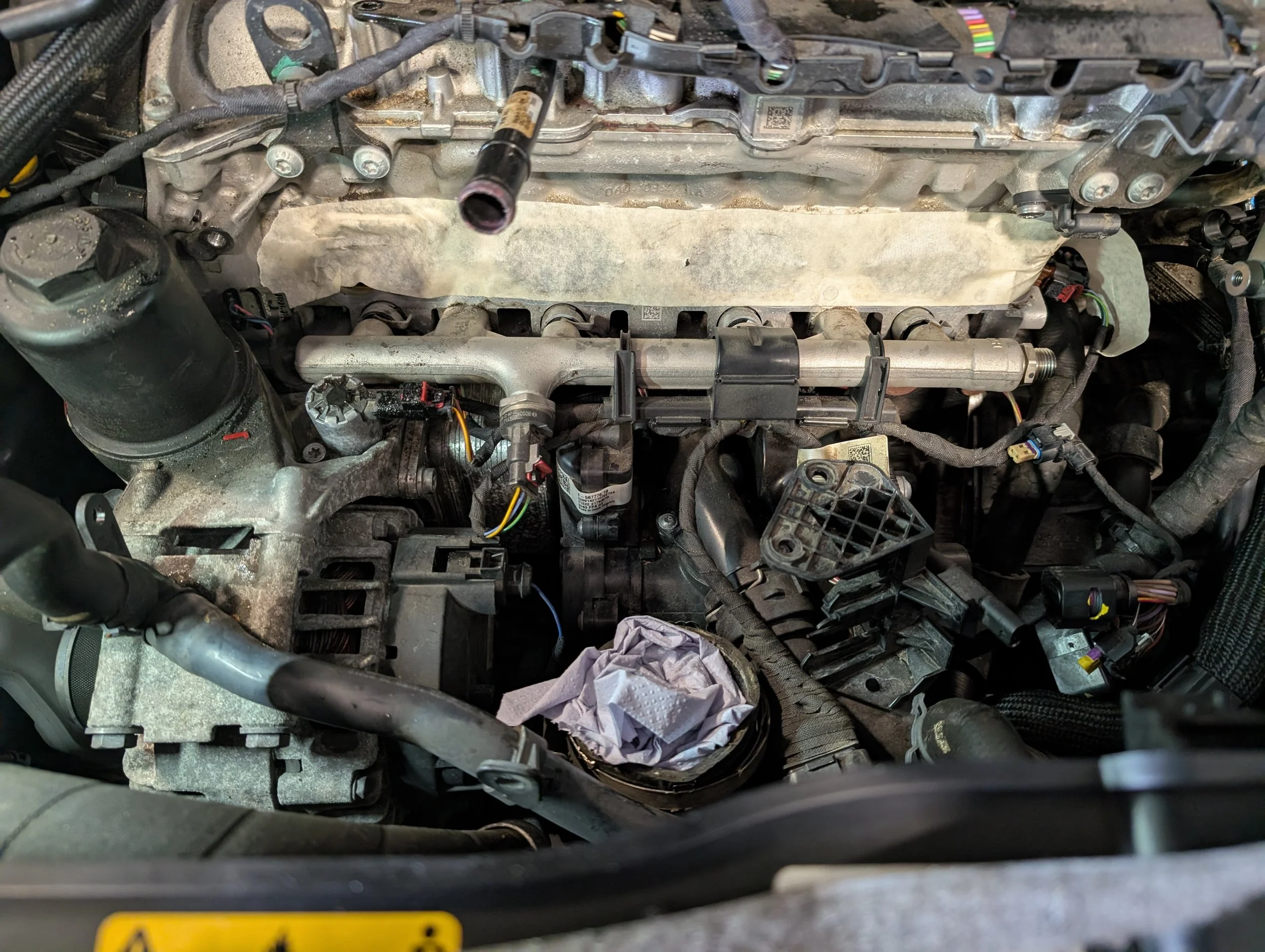 Close-up view of a car engine with various components, wires, and hoses visible. The engine appears to be undergoing repair or maintenance.