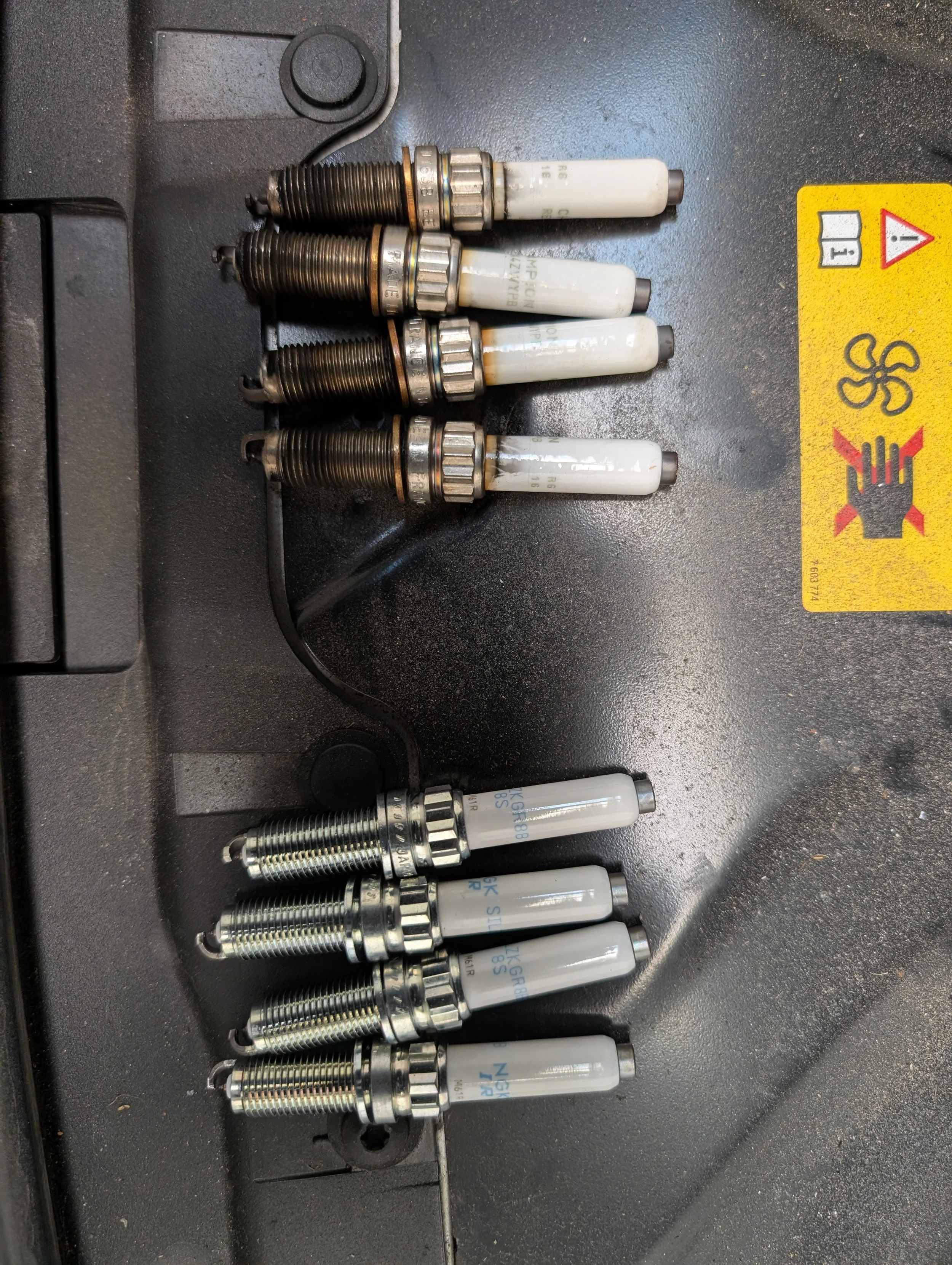 A row of spark plugs on a black surface, some are used with darkened electrodes, others are new and shiny. There is a yellow warning sticker on the right side.