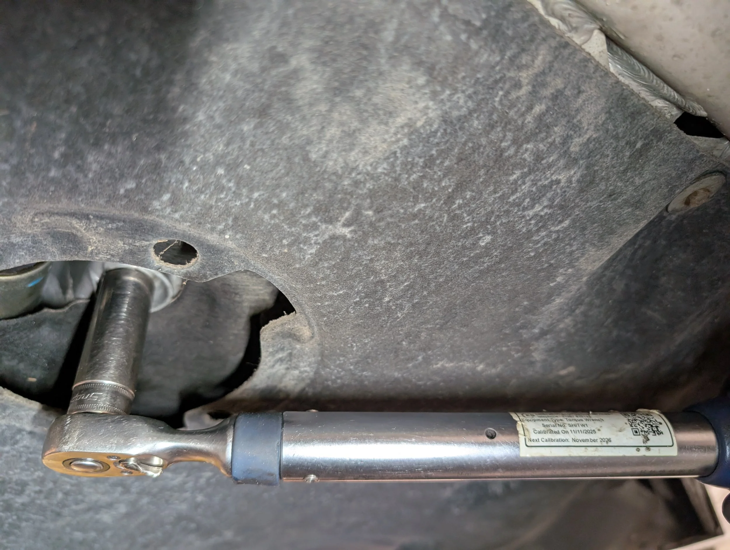 Close-up of a metallic component, possibly part of machinery or a tool, with a label containing serial number, calibration date, and QR code, attached to a textured surface.