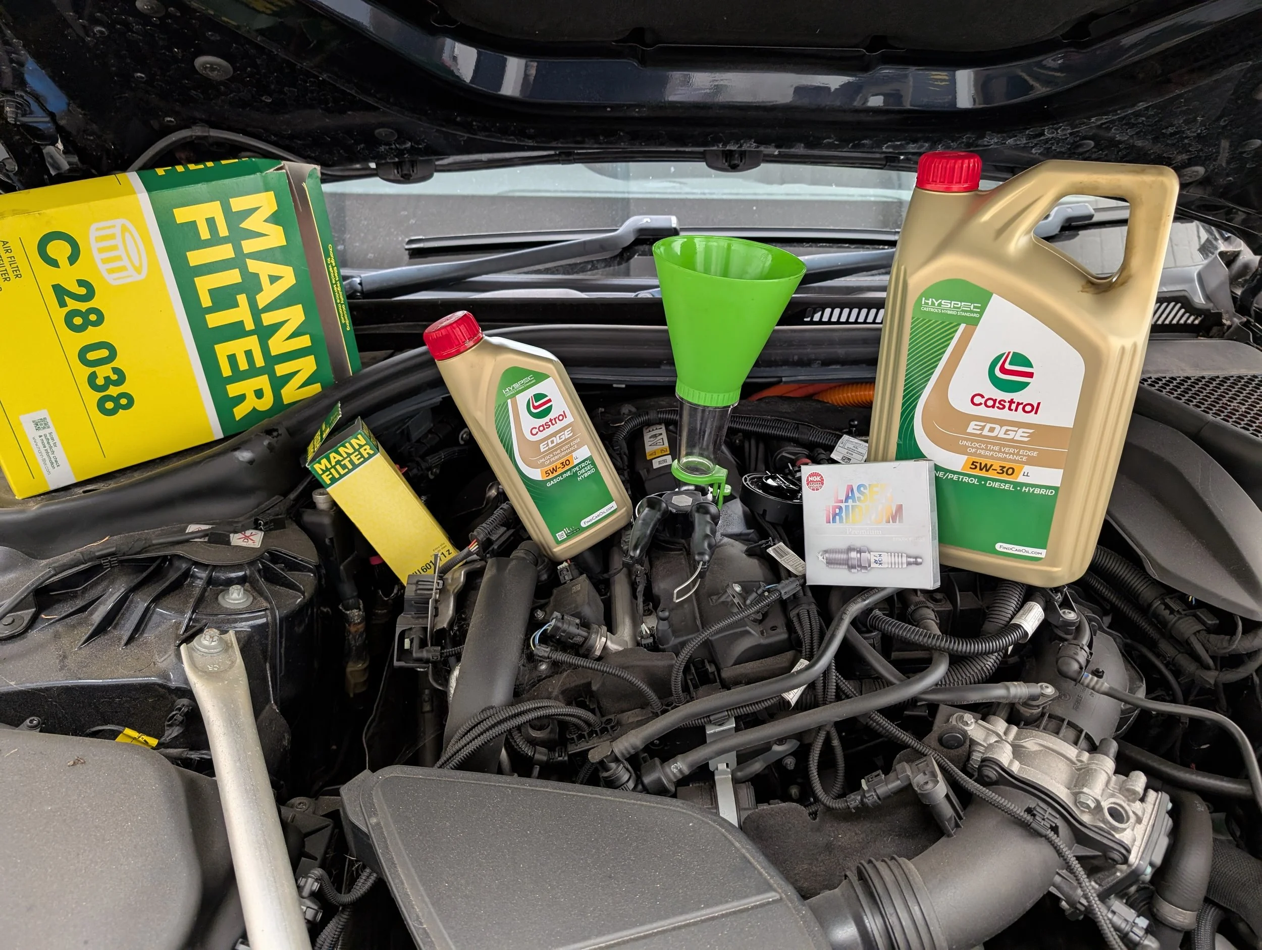 Car engine bay with motor oil, filter, and other automotive parts. The engine is partially covered with a black hood and various tools and containers are present for maintenance.