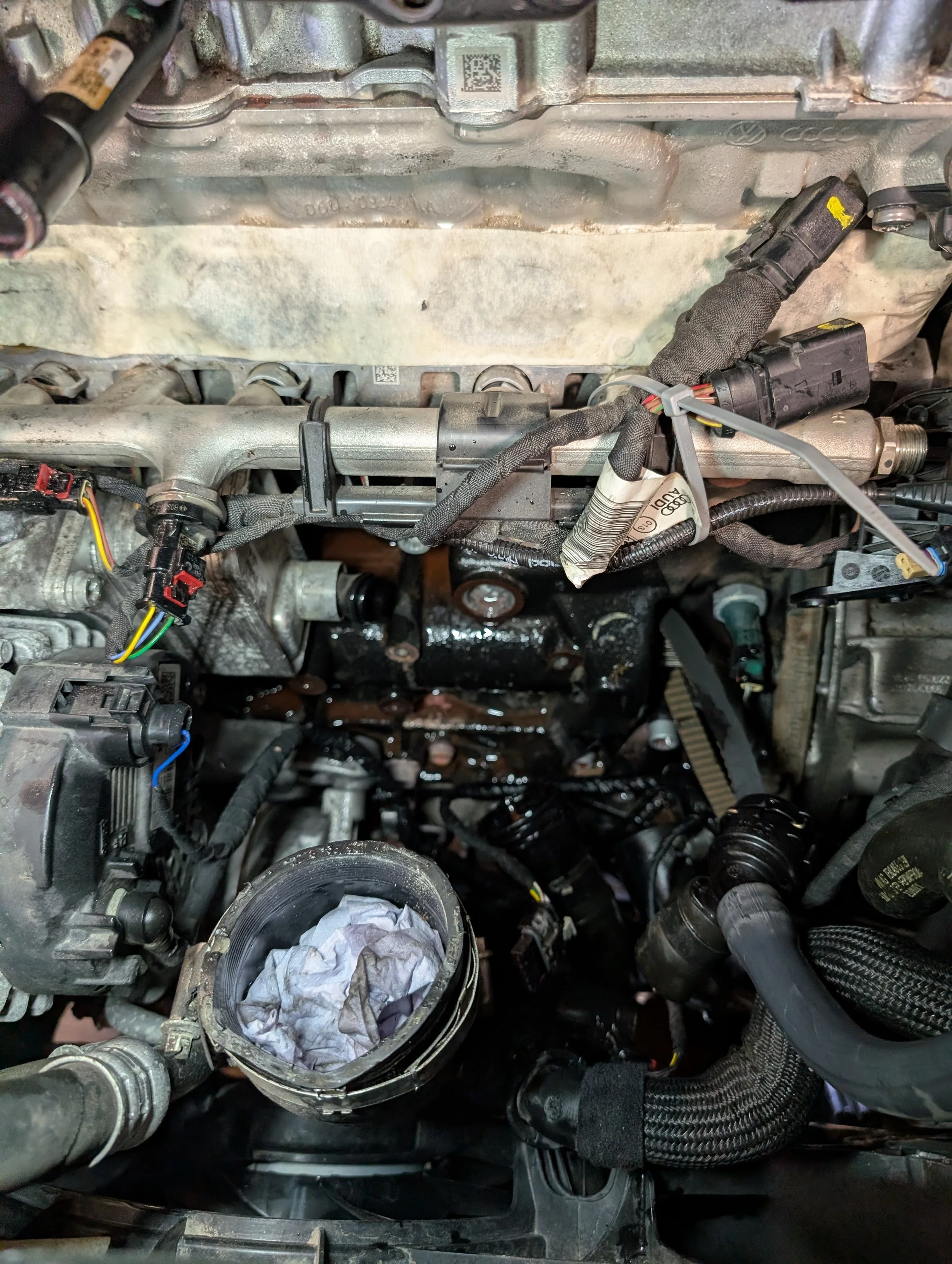 Close-up view of a disassembled car engine with various components, hoses, and wiring visible, including a cylindrical metallic part with a cloth inside.