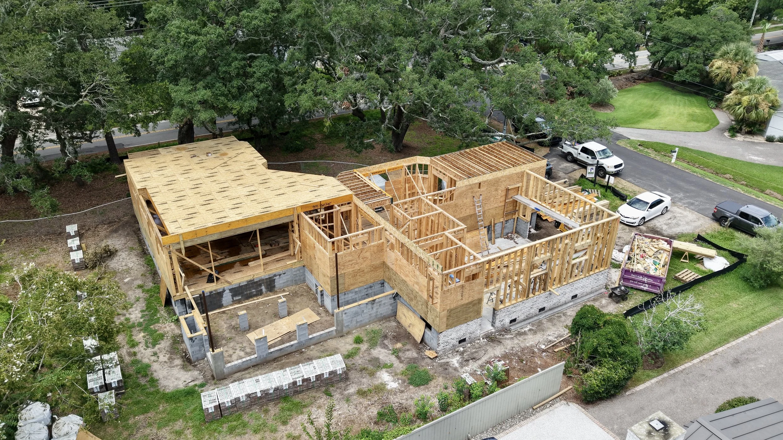 Aerial view of a house under construction, showing exposed wooden framing among surrounding trees.