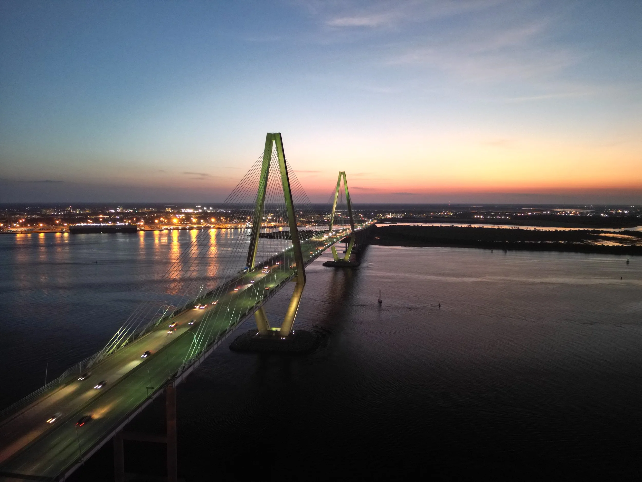 Aerial view of the Ravenel Bridge crossing Charleston Harbor at dusk, with city lights reflecting on the water.