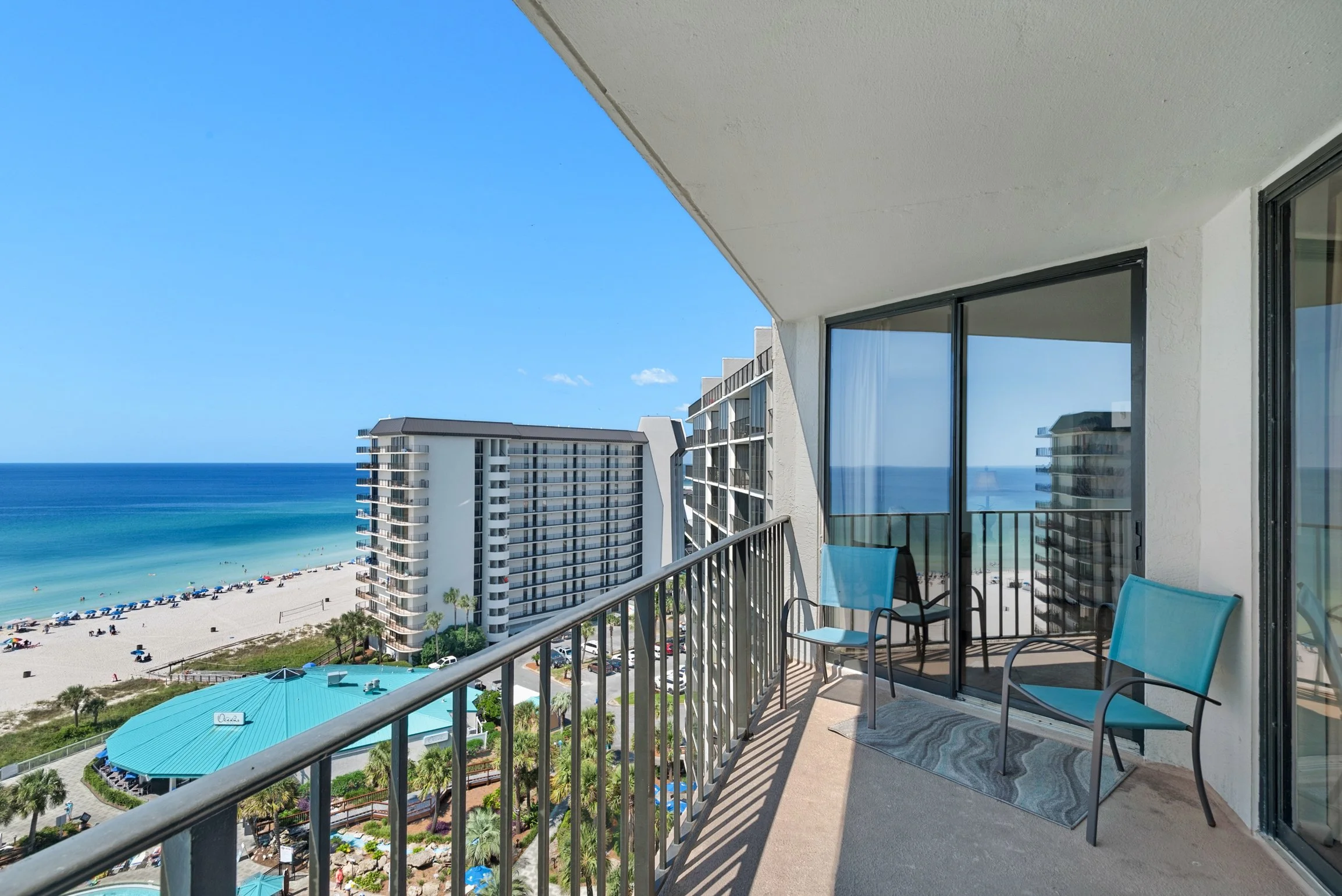 Balcony view overlooking beach and ocean, with tall white buildings, blue sky, and umbrellas on the beach.
