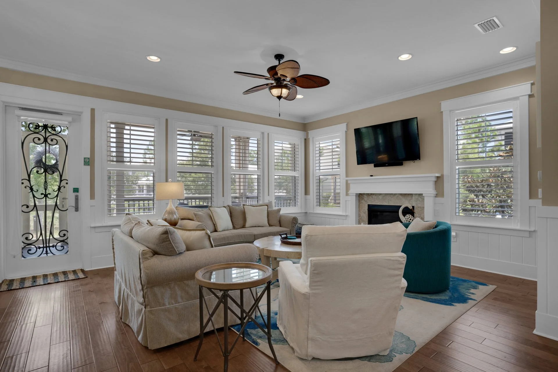Living room with beige sofas, white armchair, round wooden coffee table, large windows with white shutters, a wall-mounted TV above a fireplace, ceiling fan with wooden blades, hardwood floors, and a decorated area rug.