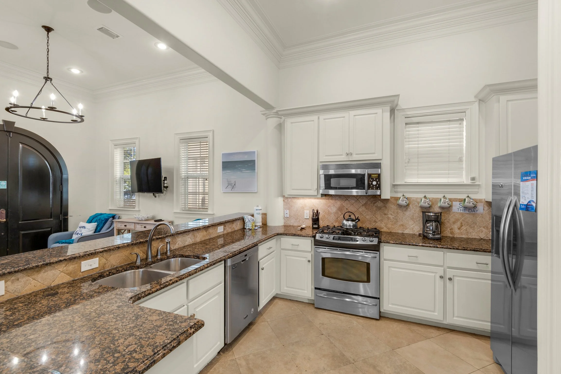 Kitchen with white cabinets, granite countertops, stainless steel oven, microwave, and refrigerator, with a TV on the wall, windows with blinds, and decorative items on the counter.