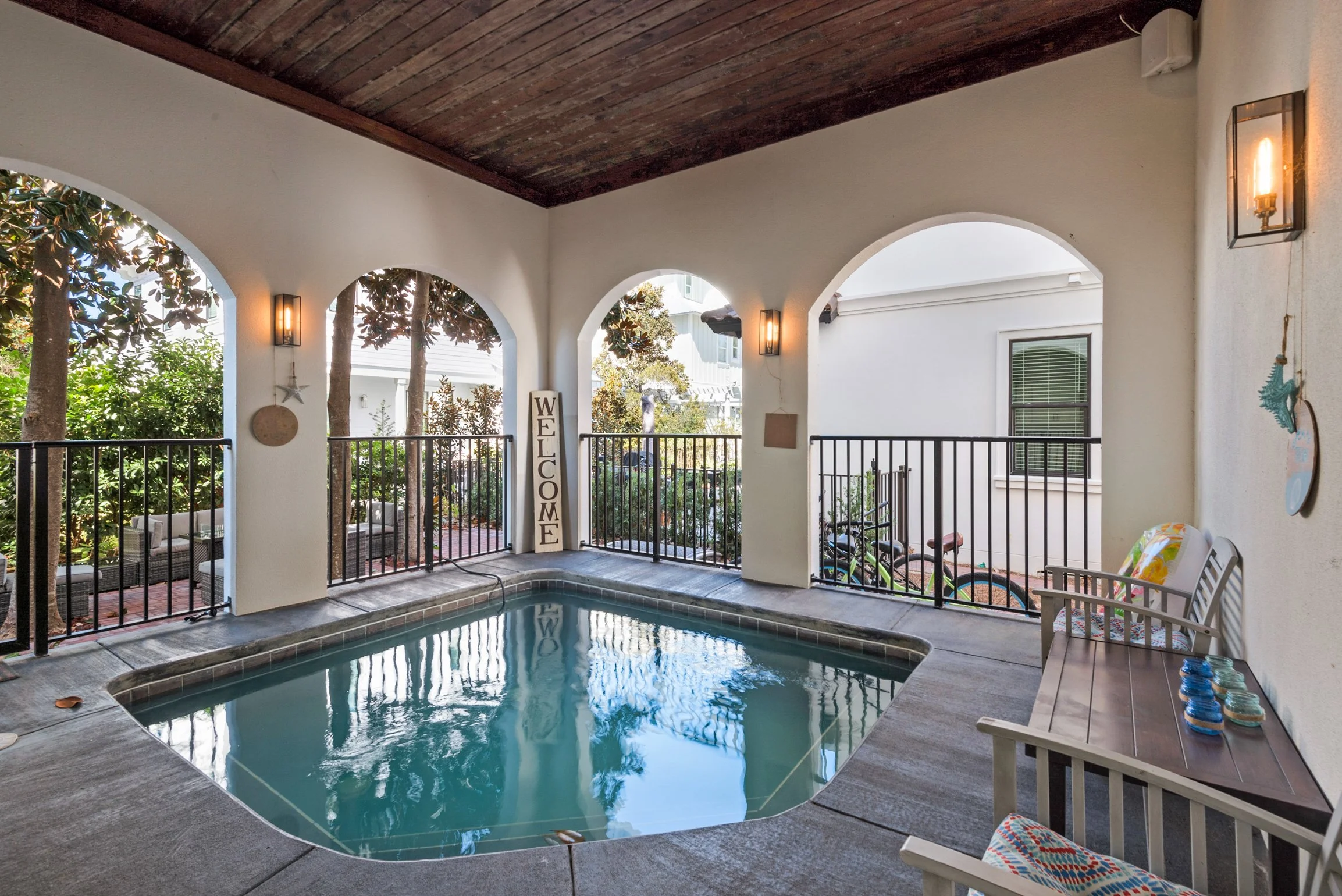 Covered porch with small swimming pool, patio furniture, and decorative items, overlooking trees and neighboring houses.