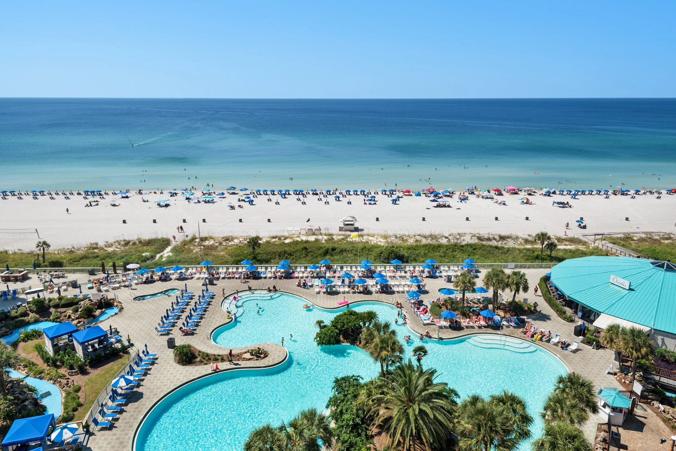 A beach scene with white sand, blue umbrellas, sunbathers, and a calm ocean with a boat. In the foreground, a swimming pool area with lounge chairs, umbrellas, and palm trees.