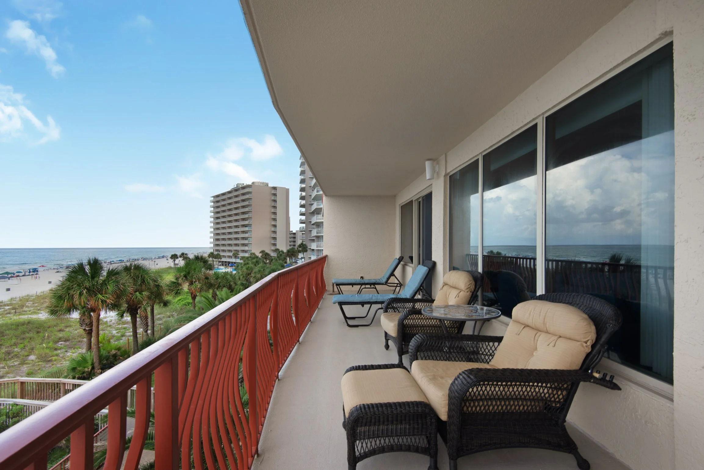 Balcony view overlooking the beach with palm trees and high-rise buildings, furnished with chairs and a table, outside a beachfront condo.