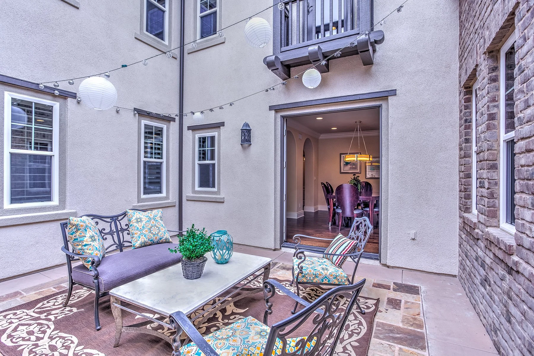 Outside patio with metal sofa and chairs with colorful cushions, a coffee table with a potted plant and lantern, string lights and paper lanterns overhead, open sliding door revealing a dining room with a wooden table and chairs, and framed pictures 