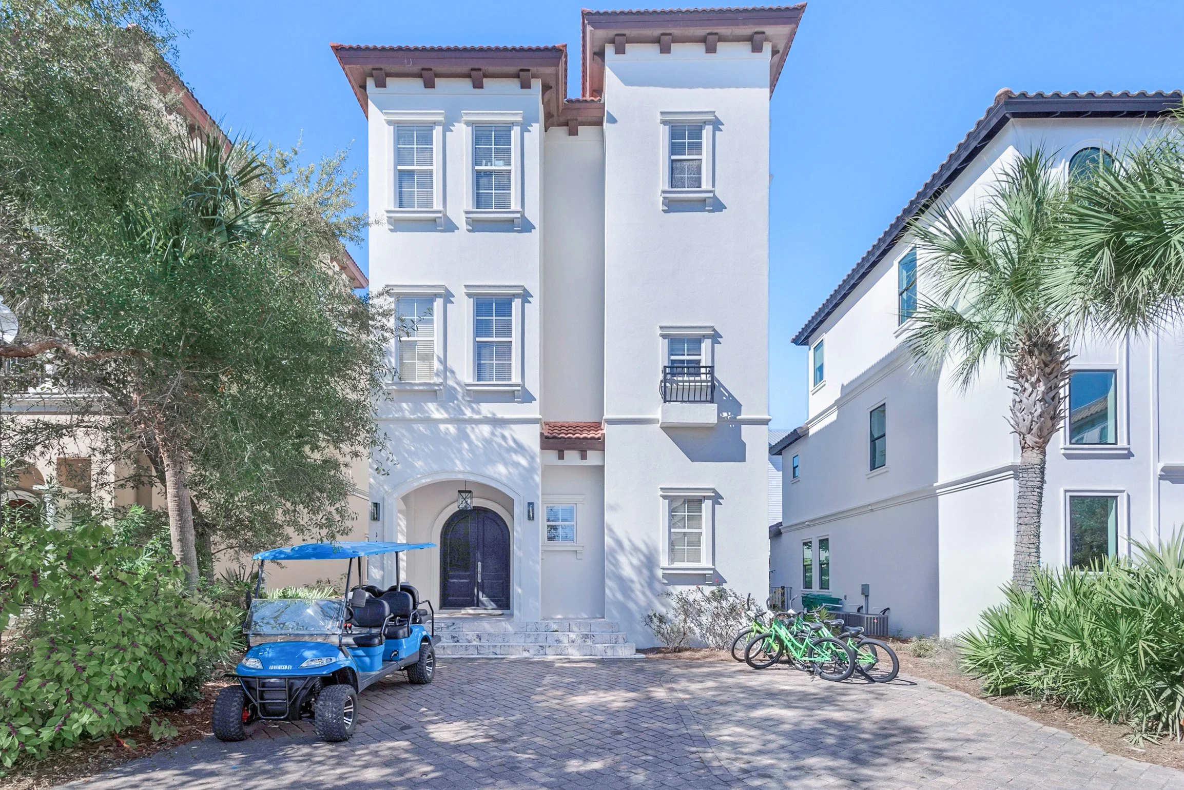 A white multi-story residential building with a black front door, surrounded by tropical plants and palm trees. A blue golf cart is parked near the entrance, and two bicycles are leaning against a tree. The area is paved with bricks and has a clear b