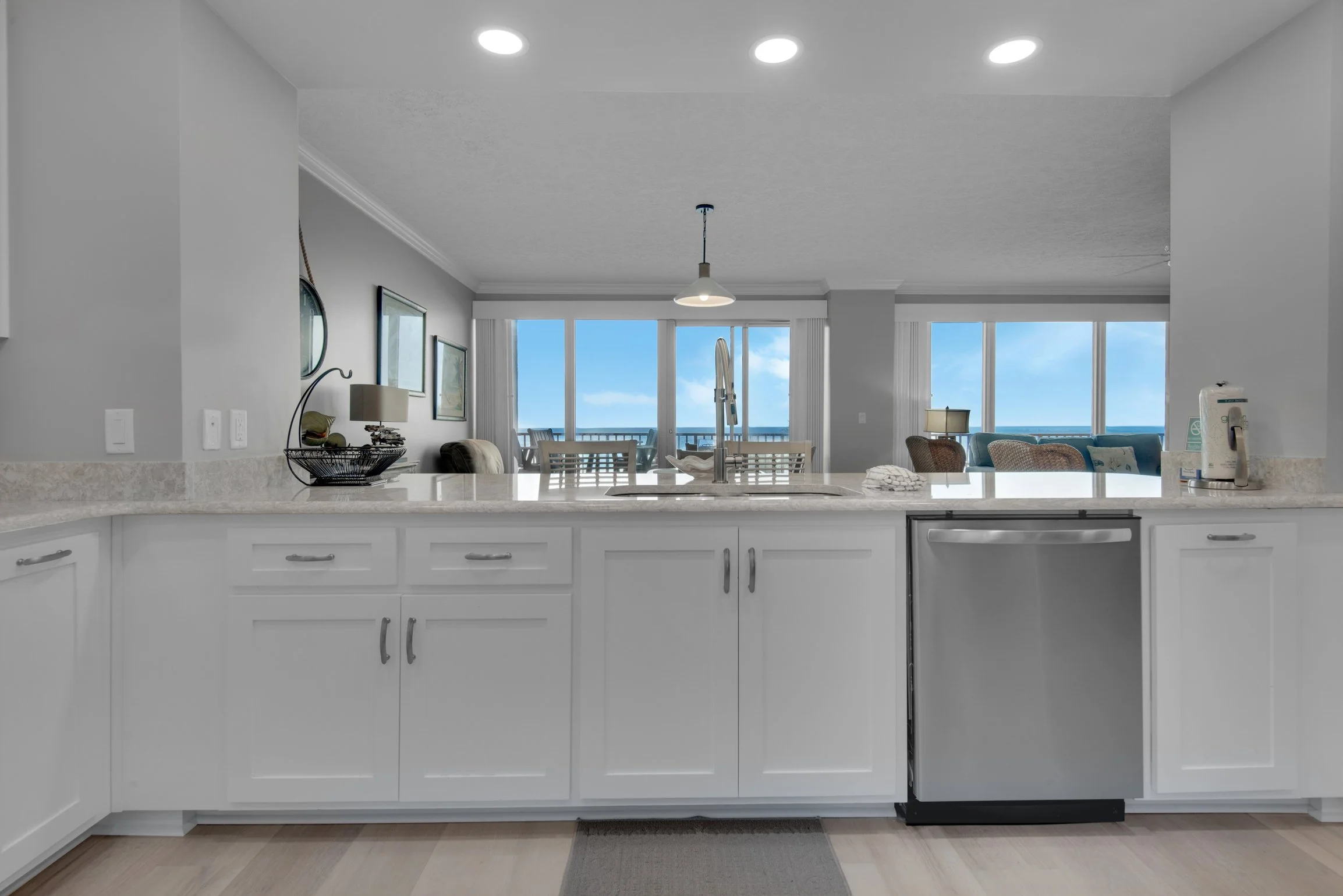 Kitchen with white cabinets, a marble countertop, a stainless steel dishwasher, and a view of a living room and balcony with ocean scenery outside.