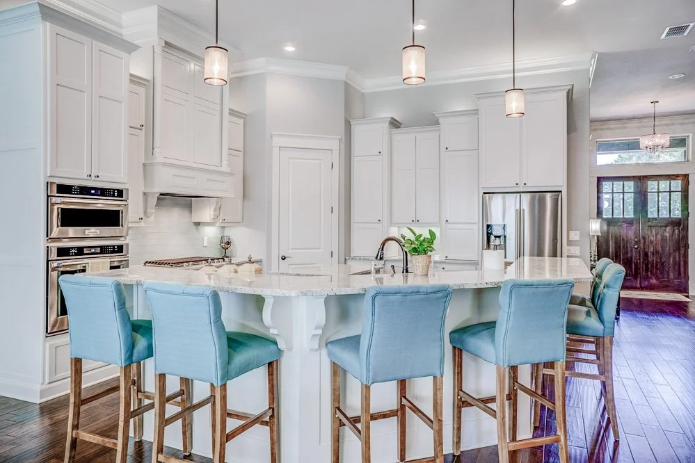 A modern kitchen with white cabinets, a large island with a granite countertop, and five blue upholstered bar stools. There are stainless steel appliances and a plant on the island. Pendant lights hang above the island.
