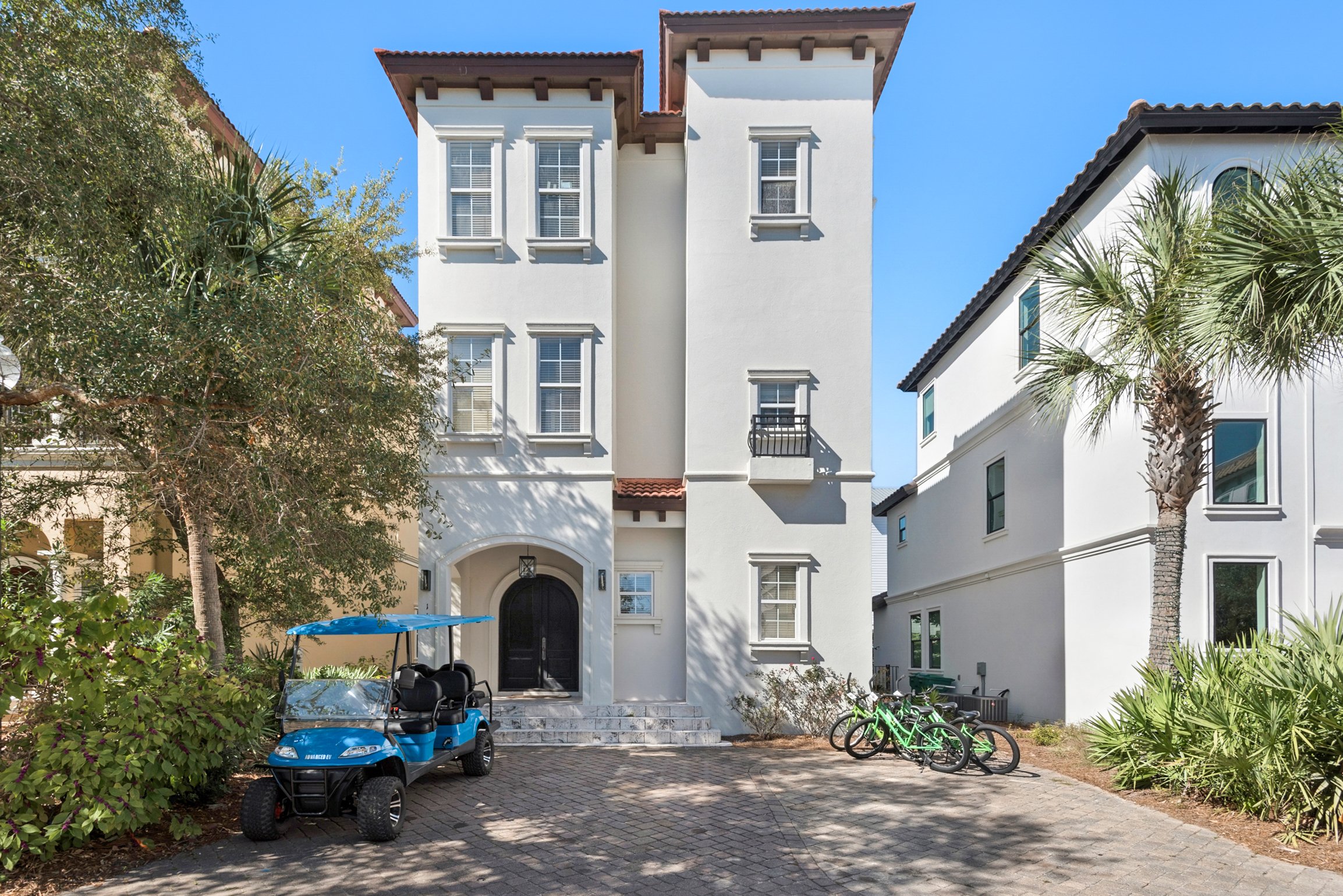 White multi-story house with multiple windows, a black front door, surrounded by trees and shrubs, with a paved driveway/parking area in front. There is a blue golf cart and several bicycles parked outside.