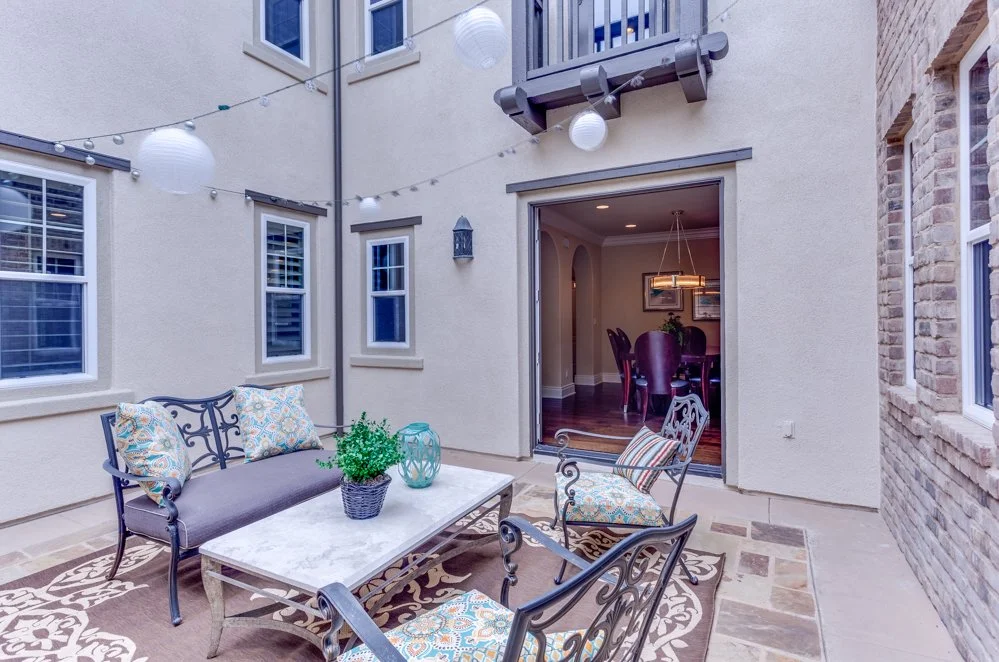 Outdoor patio area with metal furniture, decorative cushions, string lights, transitioning through a sliding door to a dining room with a table and chairs.