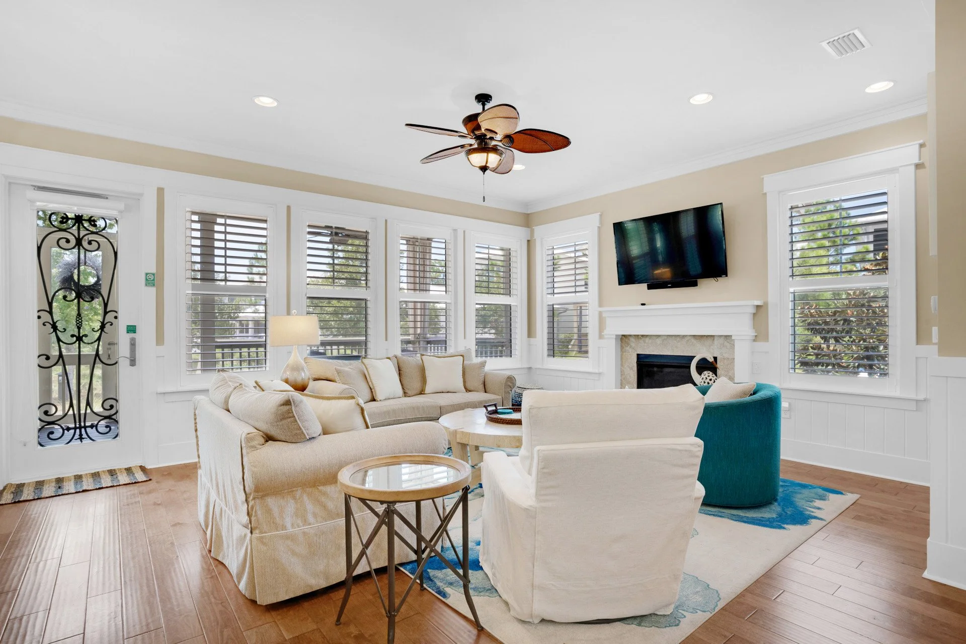 Bright living room with white walls, large windows with white blinds, beige sofa, white armchair, round coffee table, fireplace with a flat-screen TV above, ceiling fan, colorful area rug, decorative items, and wood flooring.