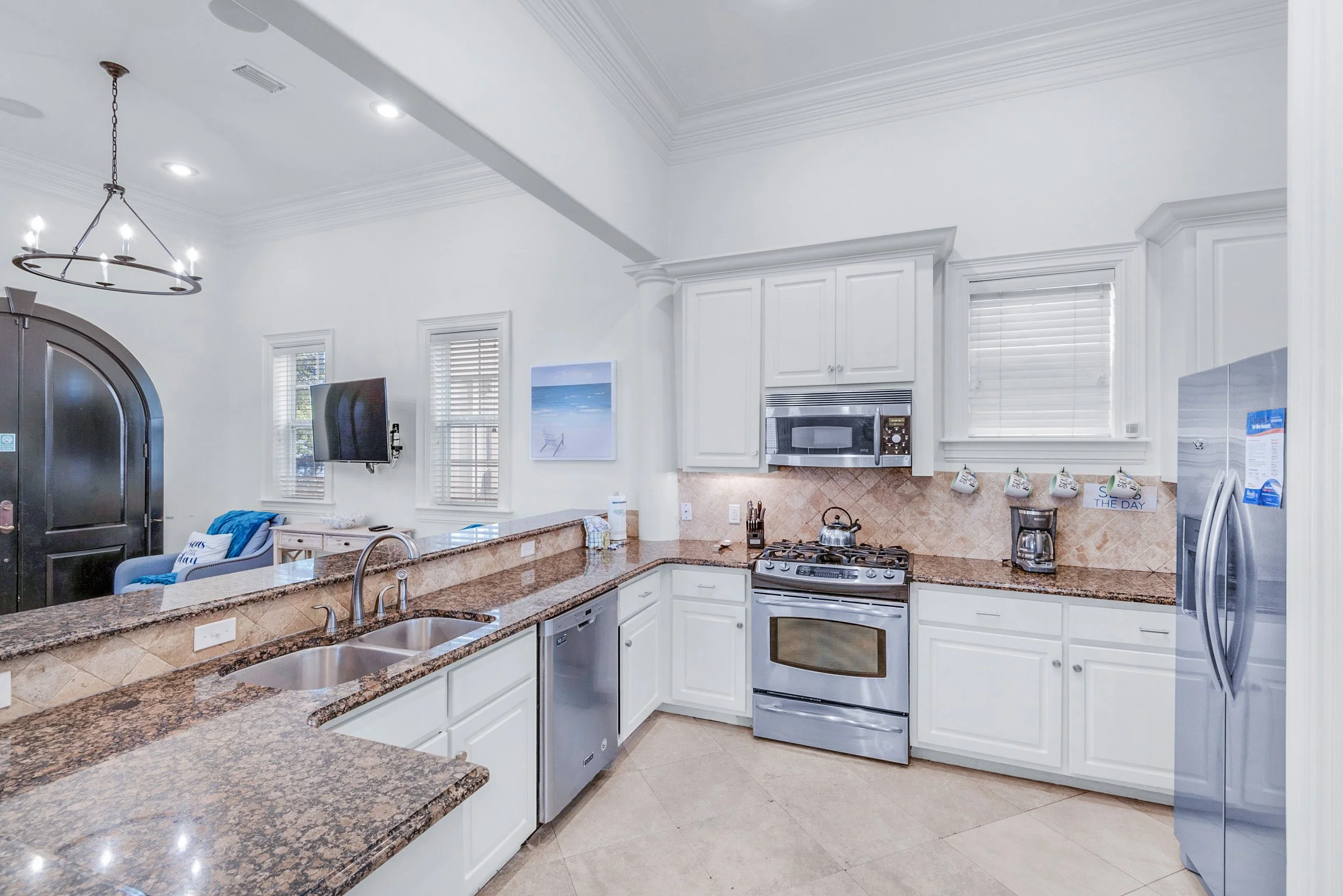 Kitchen with white cabinets, granite countertops, stainless steel appliances, a black microwave, a coffee maker, a tea kettle, and multiple decorative mugs hanging on the wall. There are windows with blinds, a wall-mounted TV, and a small painting of