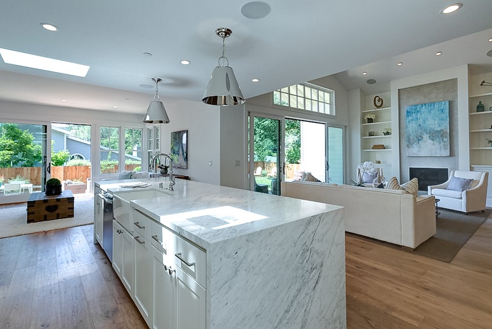Open-concept living room and kitchen with large island, white cabinetry, and minimalist decor. Sunlight streams through large windows and sliding glass doors to the backyard.
