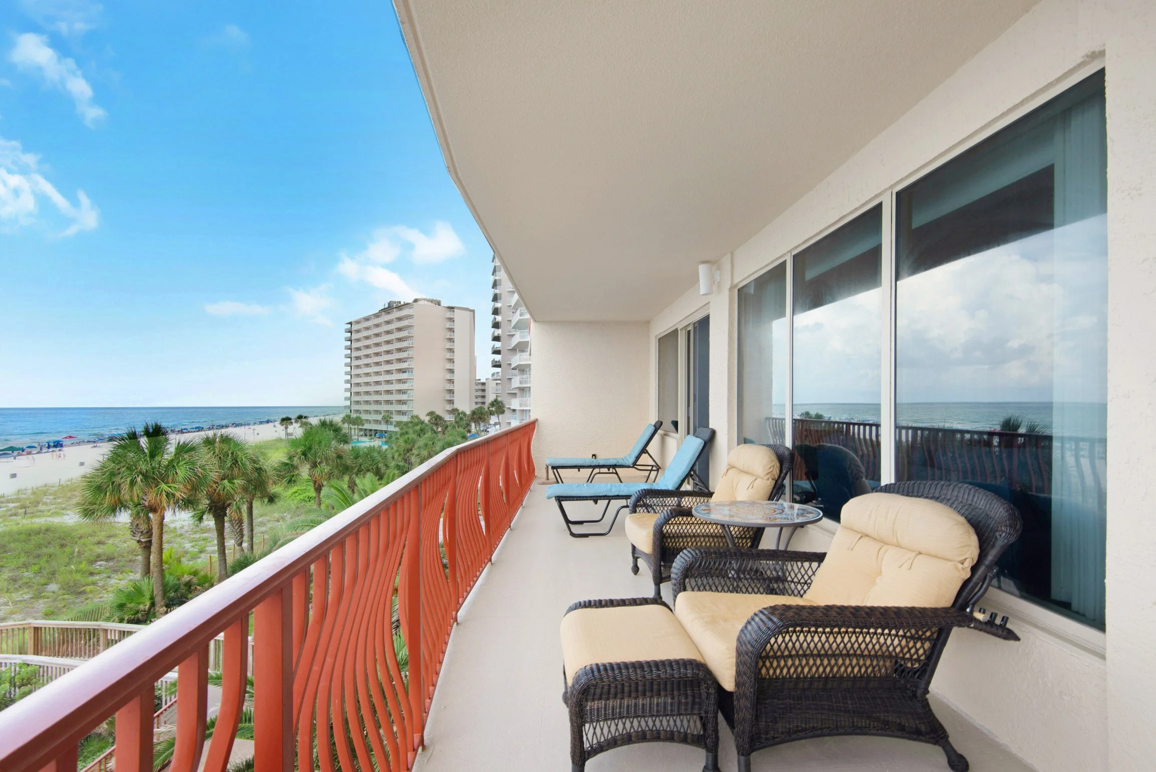 Balcony view overlooking a beach, with outdoor chairs and tables, and high-rise buildings in the distance under a partly cloudy sky.