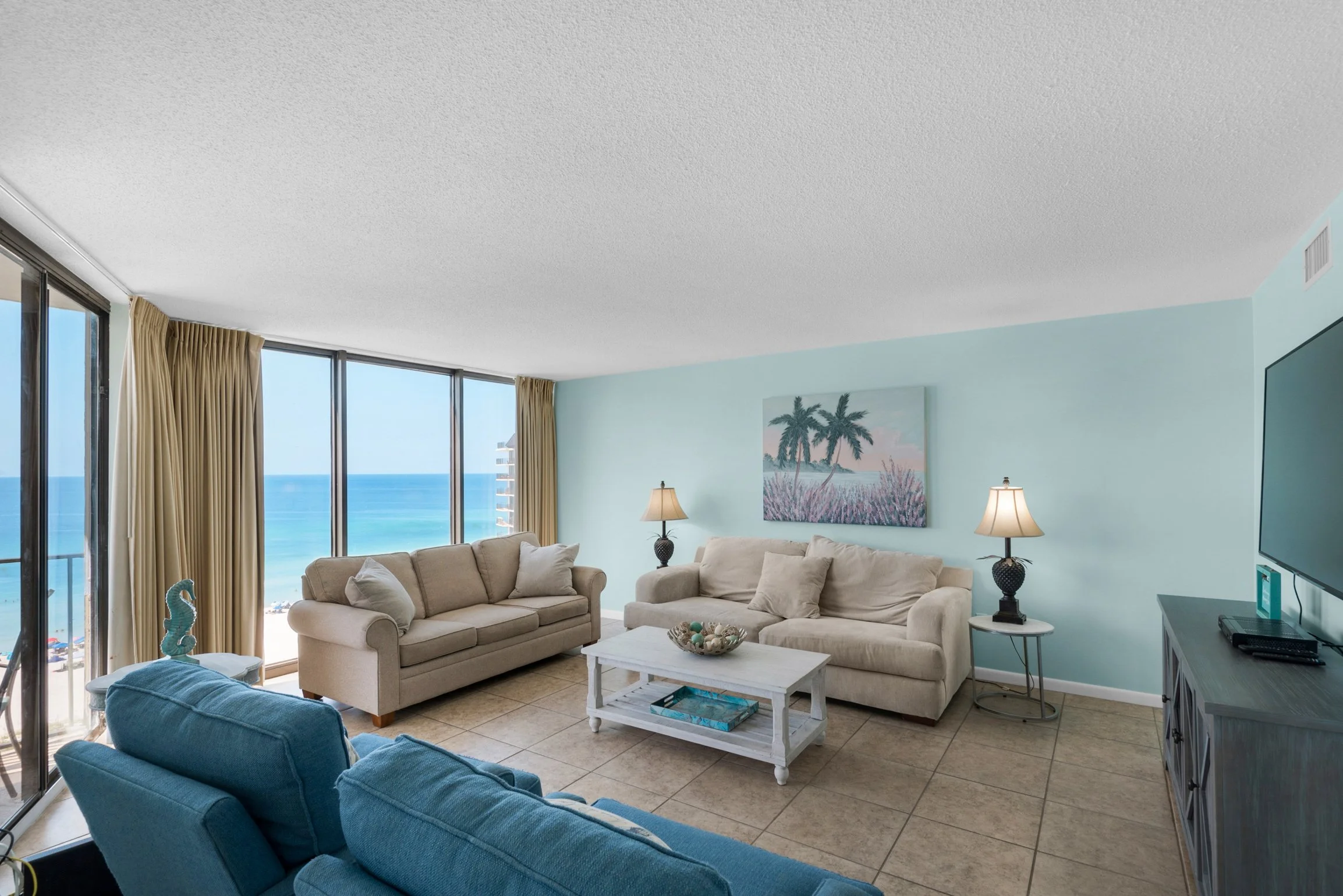 Living room with beige and blue sofas, large windows showing ocean view, wall art with palm trees, side tables with lamps, and a TV.
