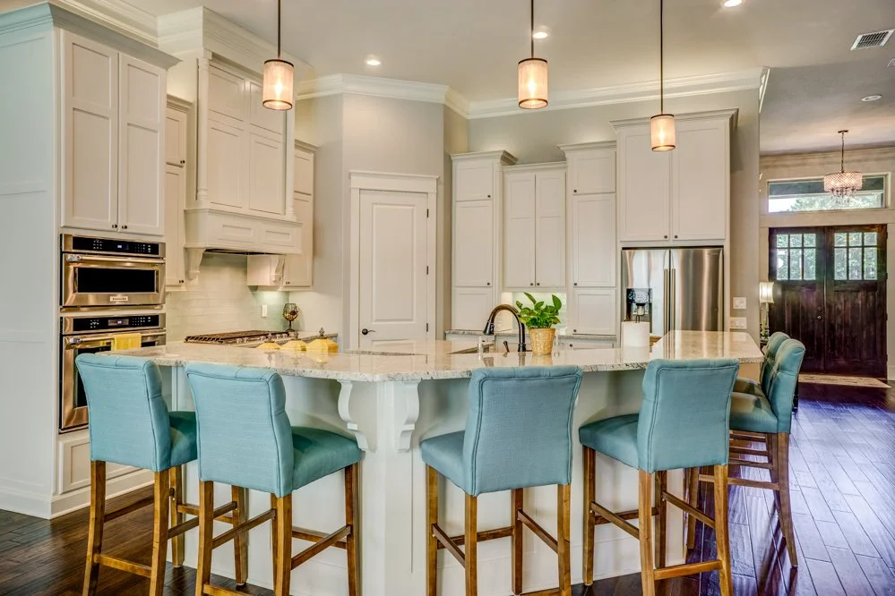 A spacious kitchen with a large white island, surrounded by six blue upholstered barstools. White cabinetry and stainless steel appliances, including double ovens and a refrigerator. Pendant lights hang above the island, and a doorway leads to a dark