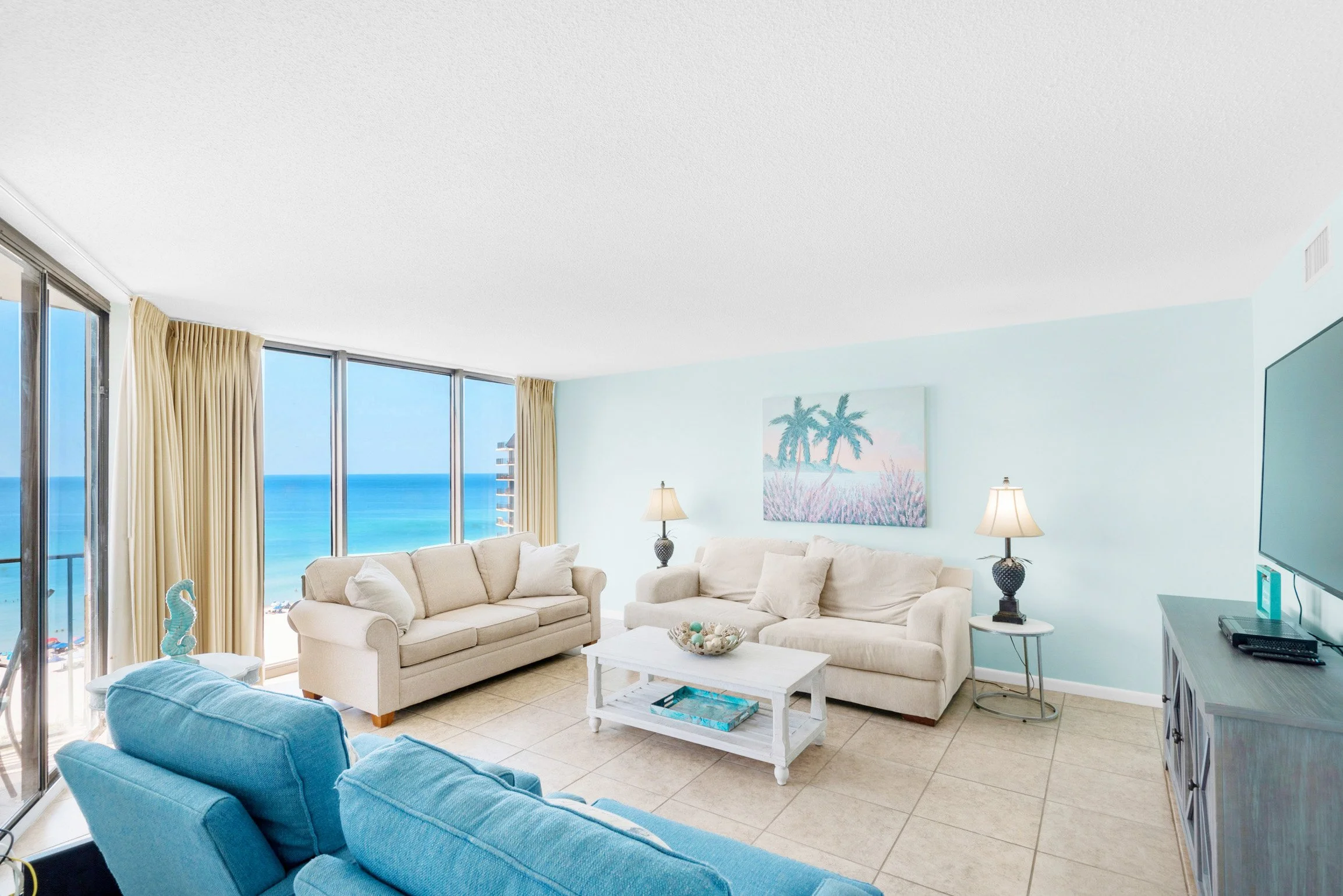 Living room with beige sofas, turquoise armchair, white coffee table, and ocean view through large windows with beige curtains.