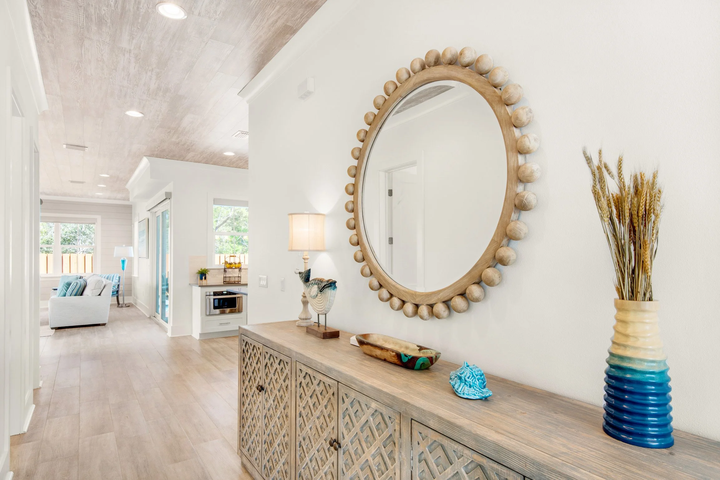 Living room with a large round mirror with ball decorations, a wooden console table with decorative objects, a vase with wheat, a lamp, windows, and light-colored wooden floors.