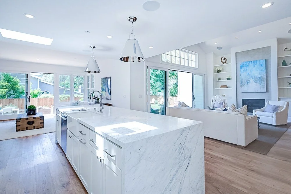 Bright open-concept living room and kitchen with white walls, large windows, and wooden flooring. The kitchen features a white marble island with pendant lighting. The living area has a white sofa, armchair, built-in shelves, and a fireplace. Sliding