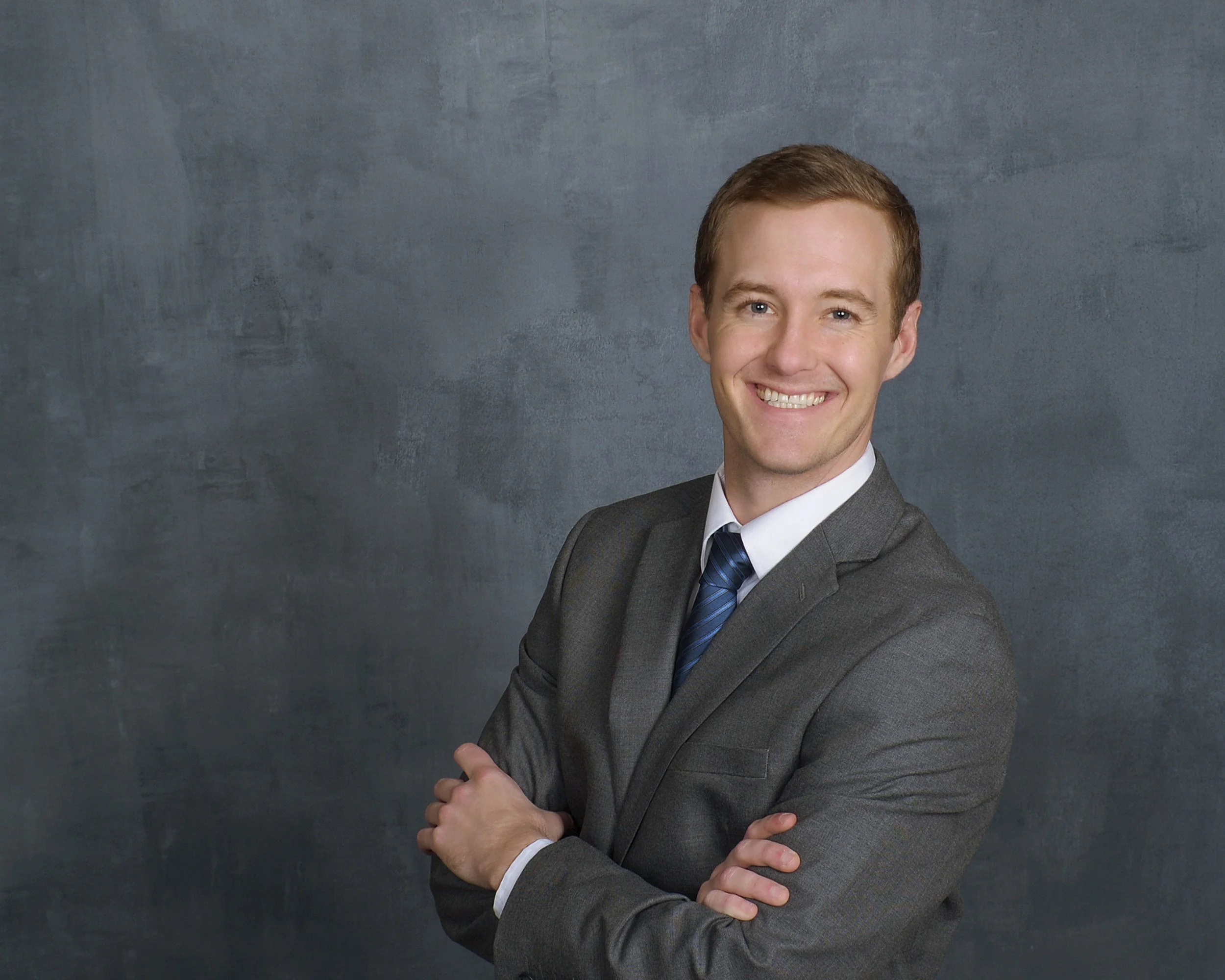 A young Caucasian man with short light brown hair, wearing a gray suit, white shirt, and blue striped tie, smiling and crossing his arms, standing against a textured gray background.