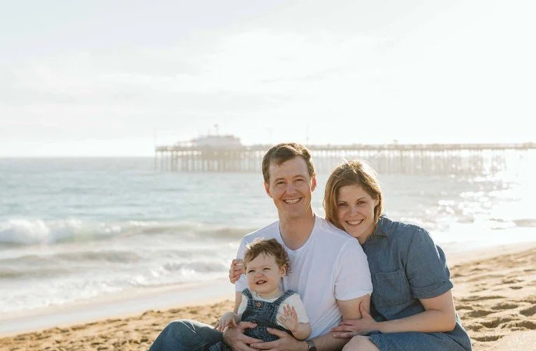 A family of three sitting on a beach, smiling at the camera with a pier in the background during sunset.