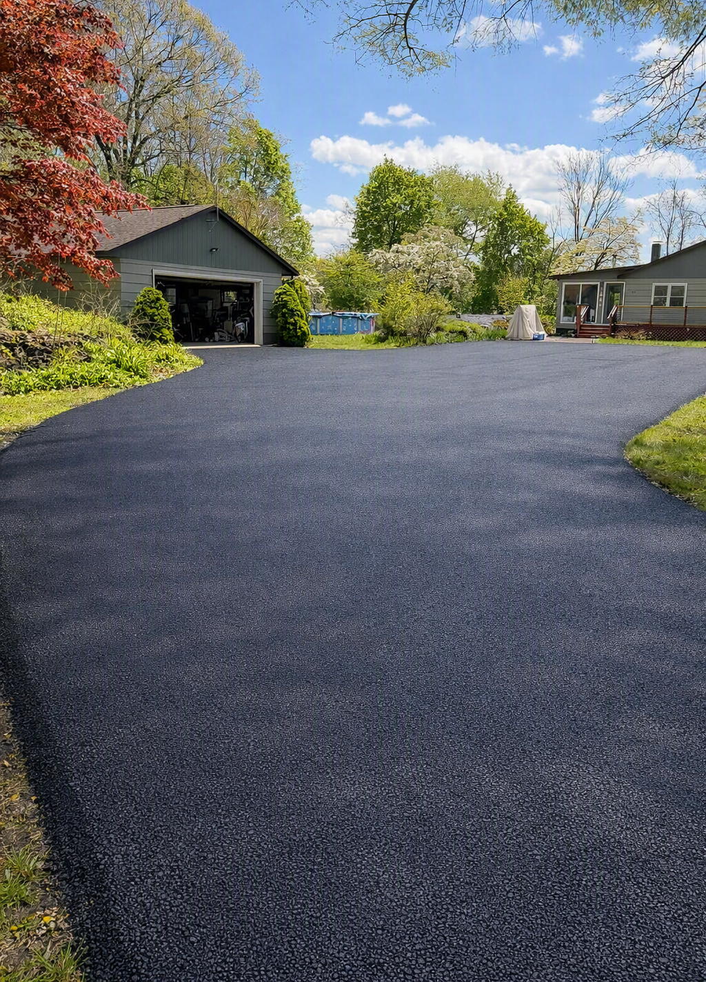 Newly paved black asphalt driveway leading to a house and garage, with trees, grass, and blue sky in the background.