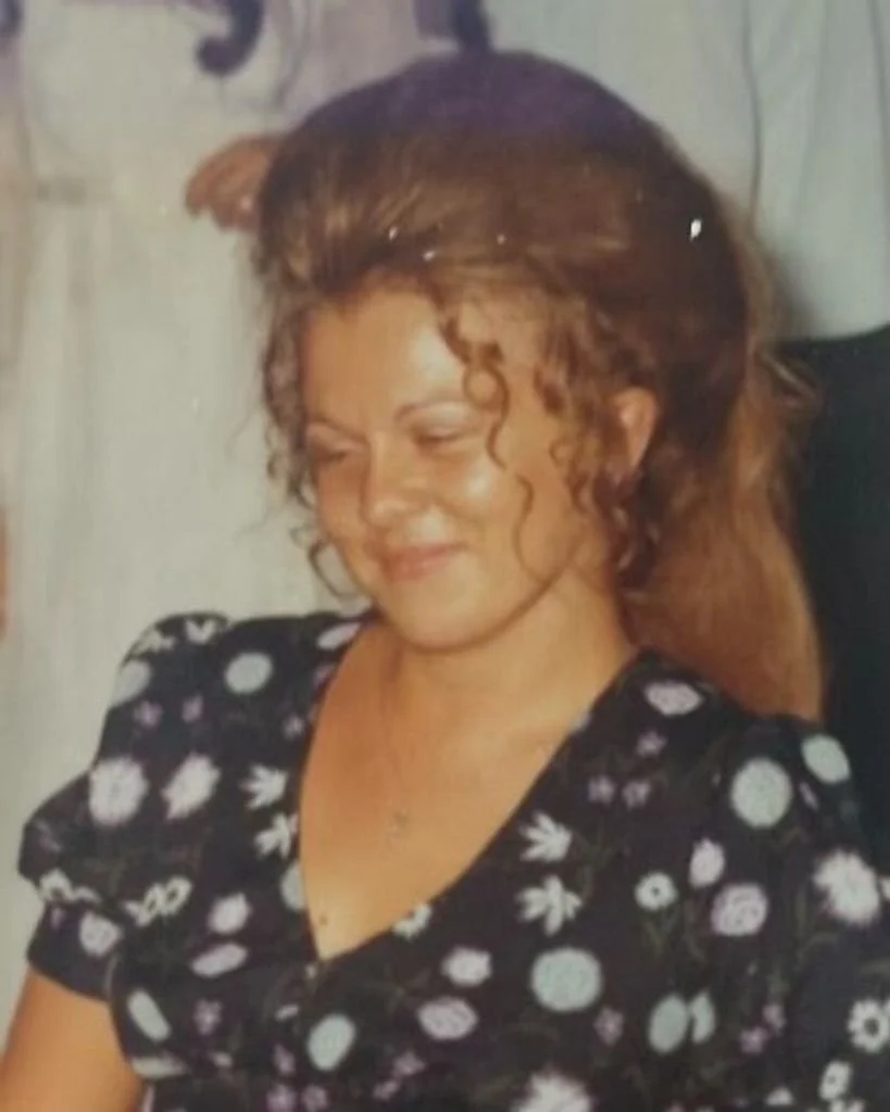 A woman with curly brown hair smiling with her eyes closed, wearing a black dress with white floral patterns.