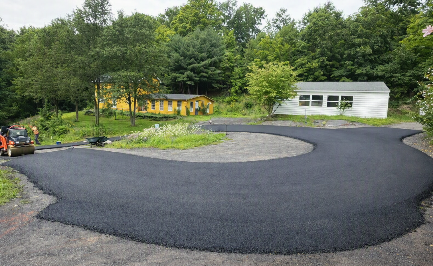 A freshly paved circular driveway in a residential area with houses and trees in the background.