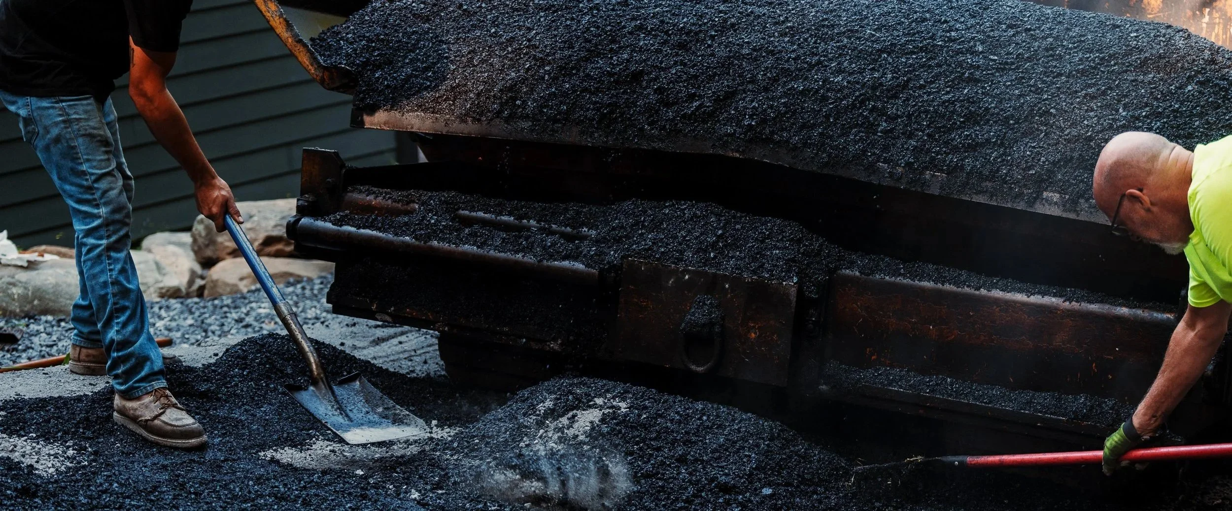 Two men are working on asphalt paving; one man is shoveling hot black asphalt while the other is using a tool to spread the asphalt on the ground.