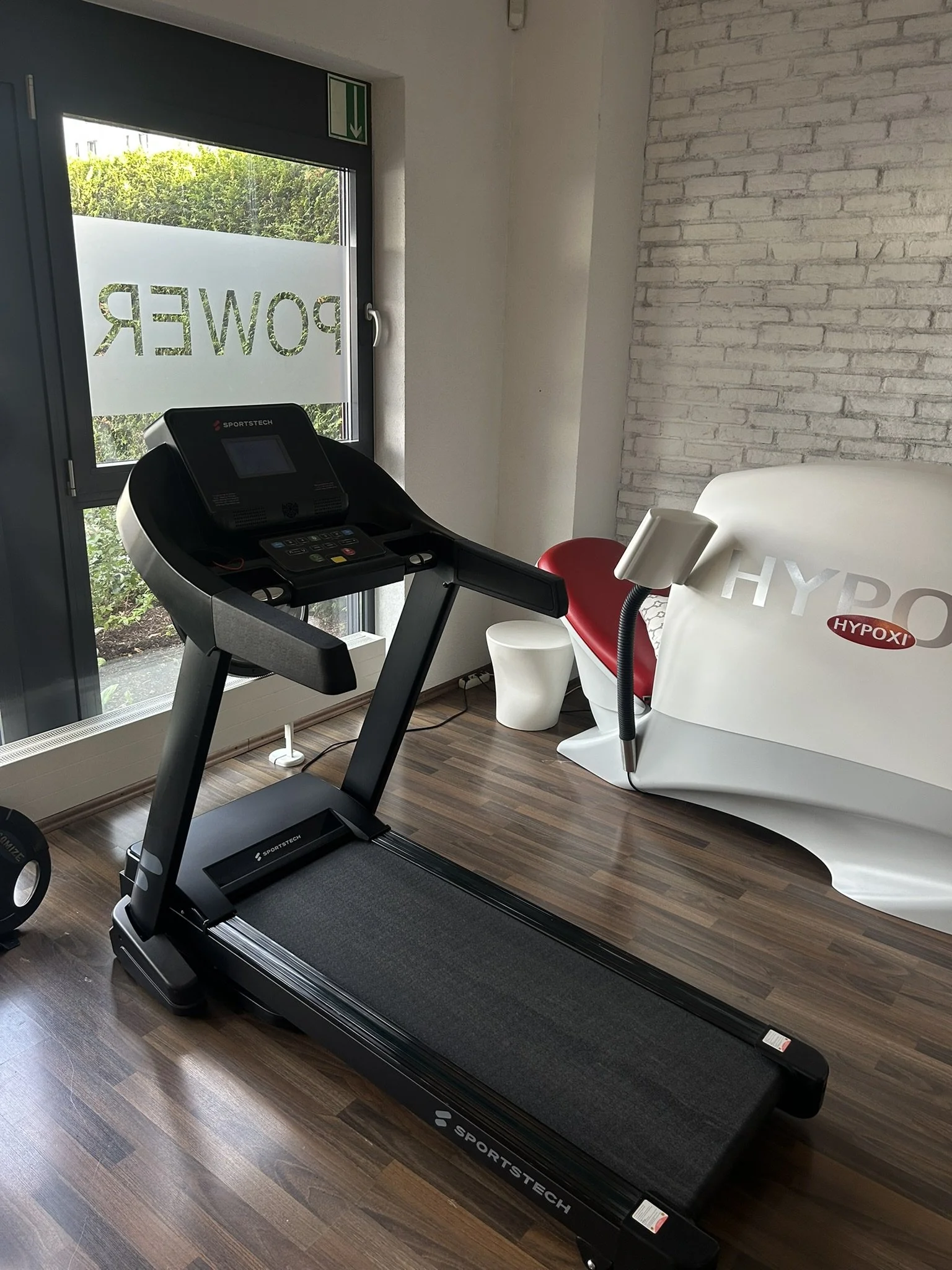 Ein Laufband im Fitnessstudio mit einer weißen Liege und einem roten Stuhl davor, an eine weiße Wand mit Ziegelmuster. Ein Fenster mit der Aufschrift 'POWER' im Hintergrund.