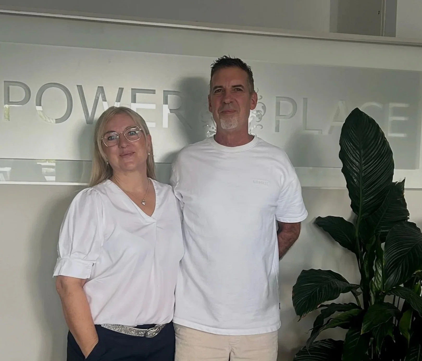 Zwei Menschen stehen vor einem weißen Hintergrund mit dem Worten "Power Place". Eine Frau mit blonde Haare, Brille und weißem Kleid, ein Mann mit dunklem Haar, Bart und weißem T-Shirt. Ein grüner Pflanzenstrauch steht rechts.