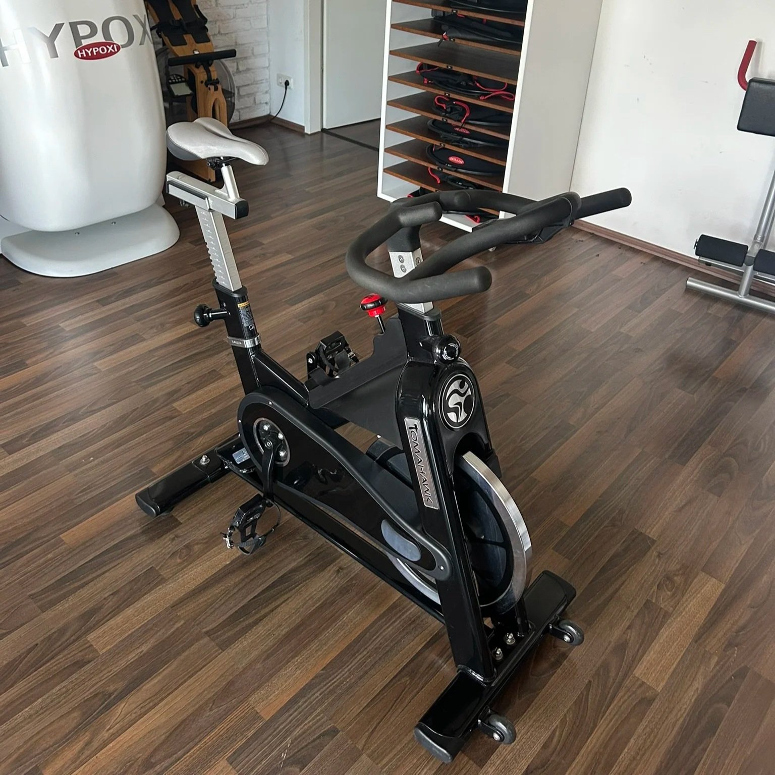 Ein Heimtrainer in einem Fitnessraum mit Holzfußboden, im Hintergrund eine Wand mit einem Regal und Fitnessgeräten, und eine elektrische Anschlussdose